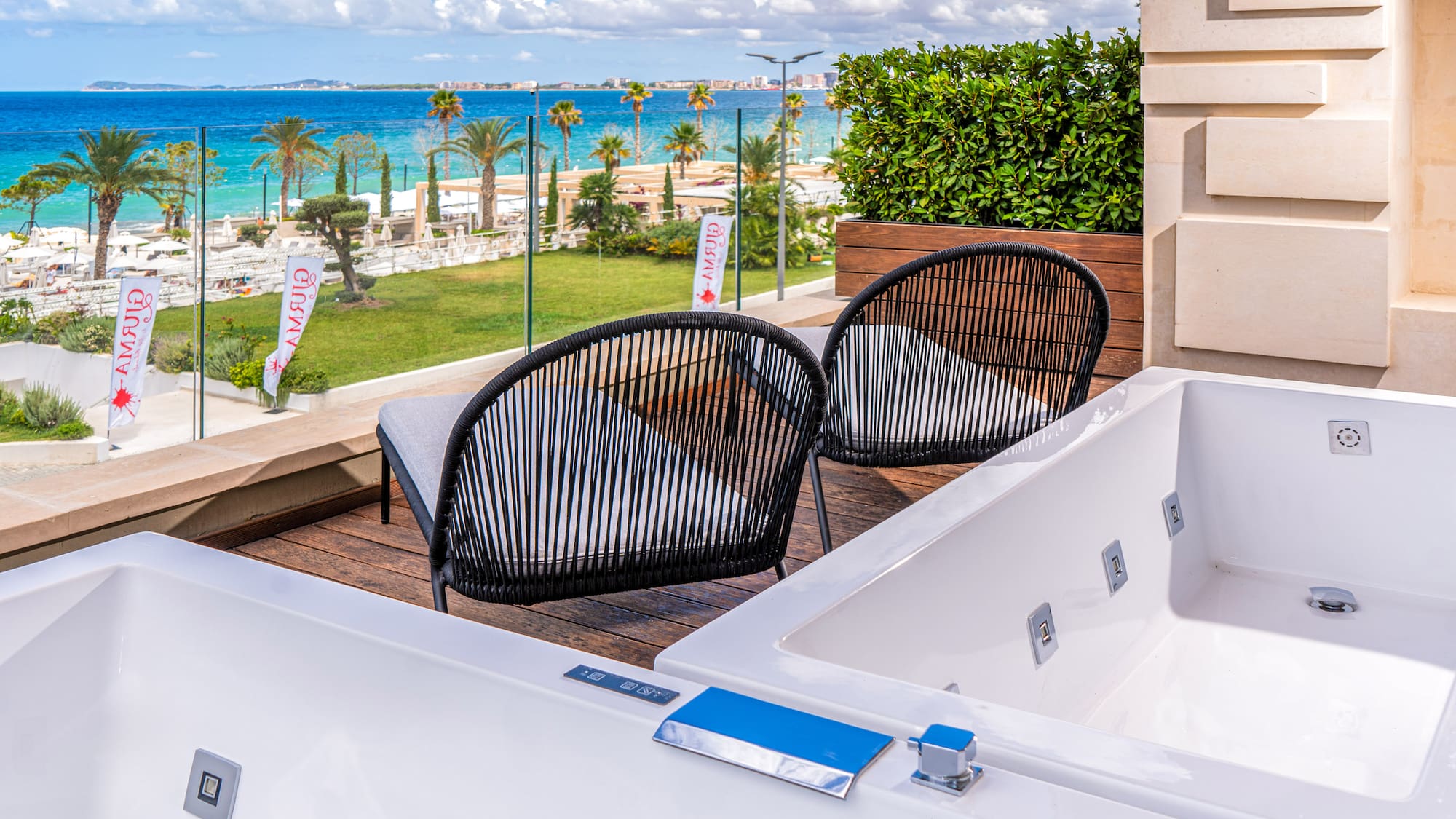 Resort balcony with two outdoor jacuzzis, chairs, beach, and blue sea view.