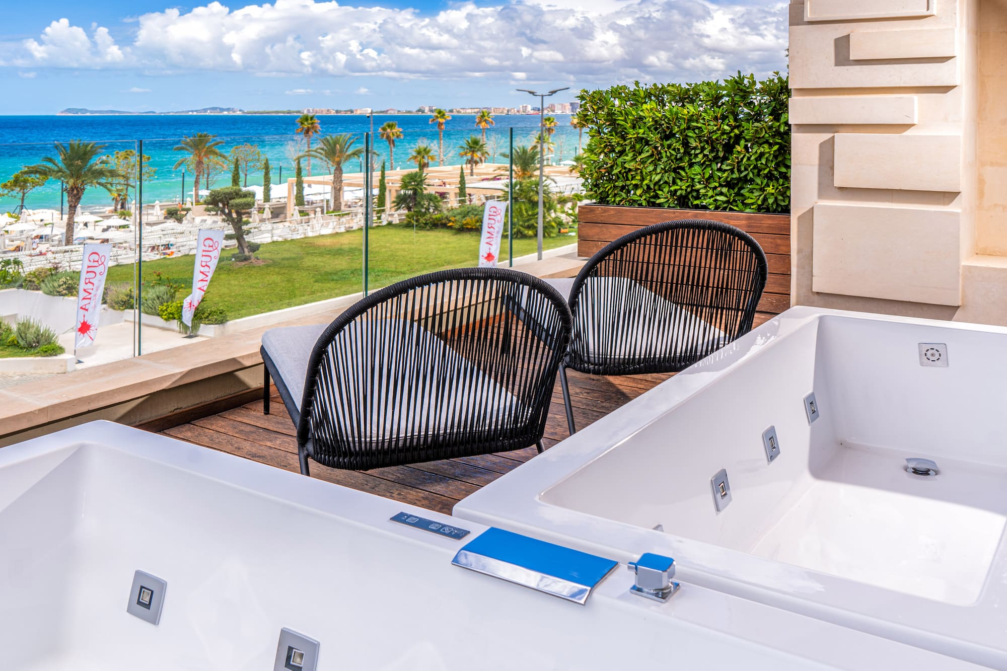 Resort balcony with two outdoor jacuzzis, chairs, beach, and blue sea view.