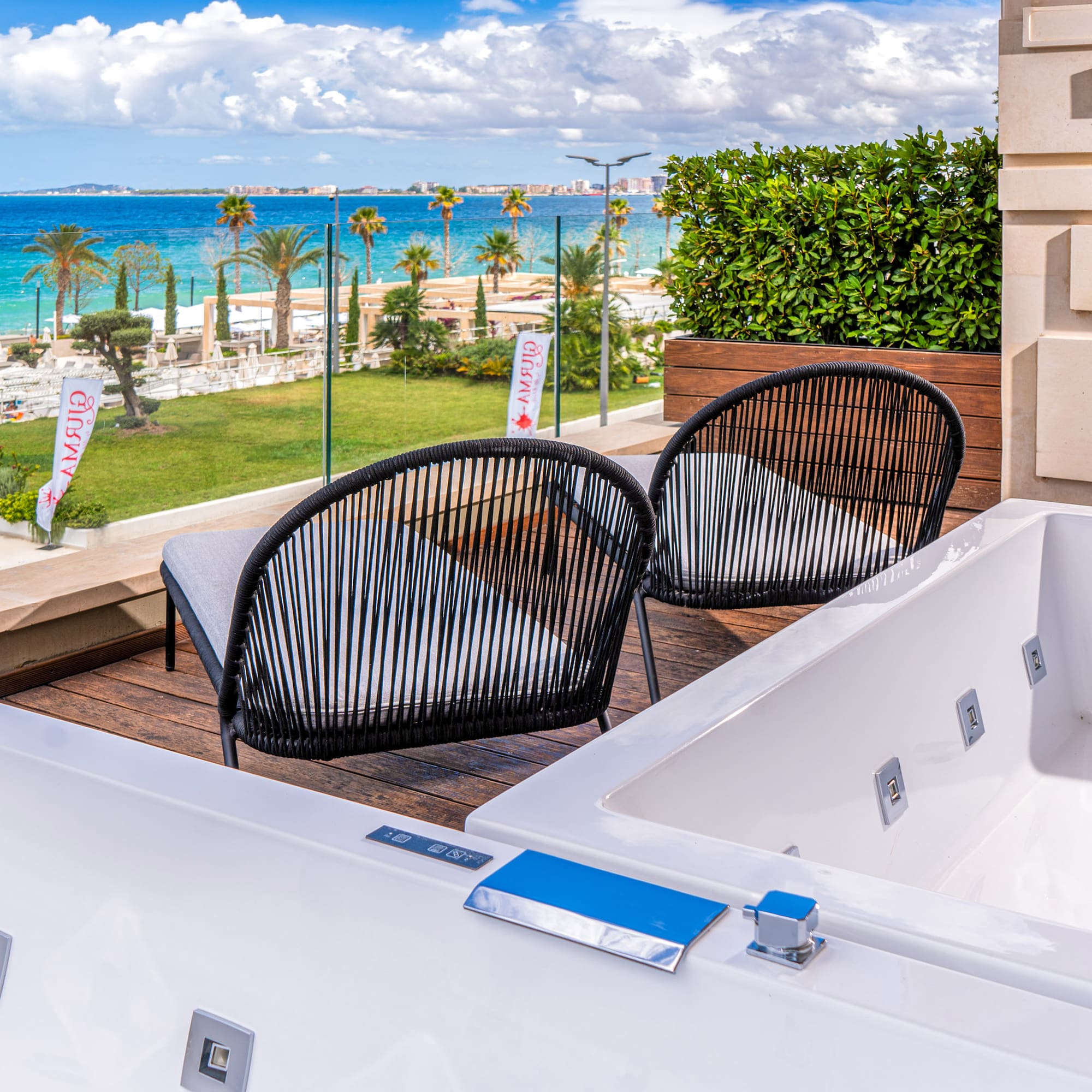 Resort balcony with two outdoor jacuzzis, chairs, beach, and blue sea view.