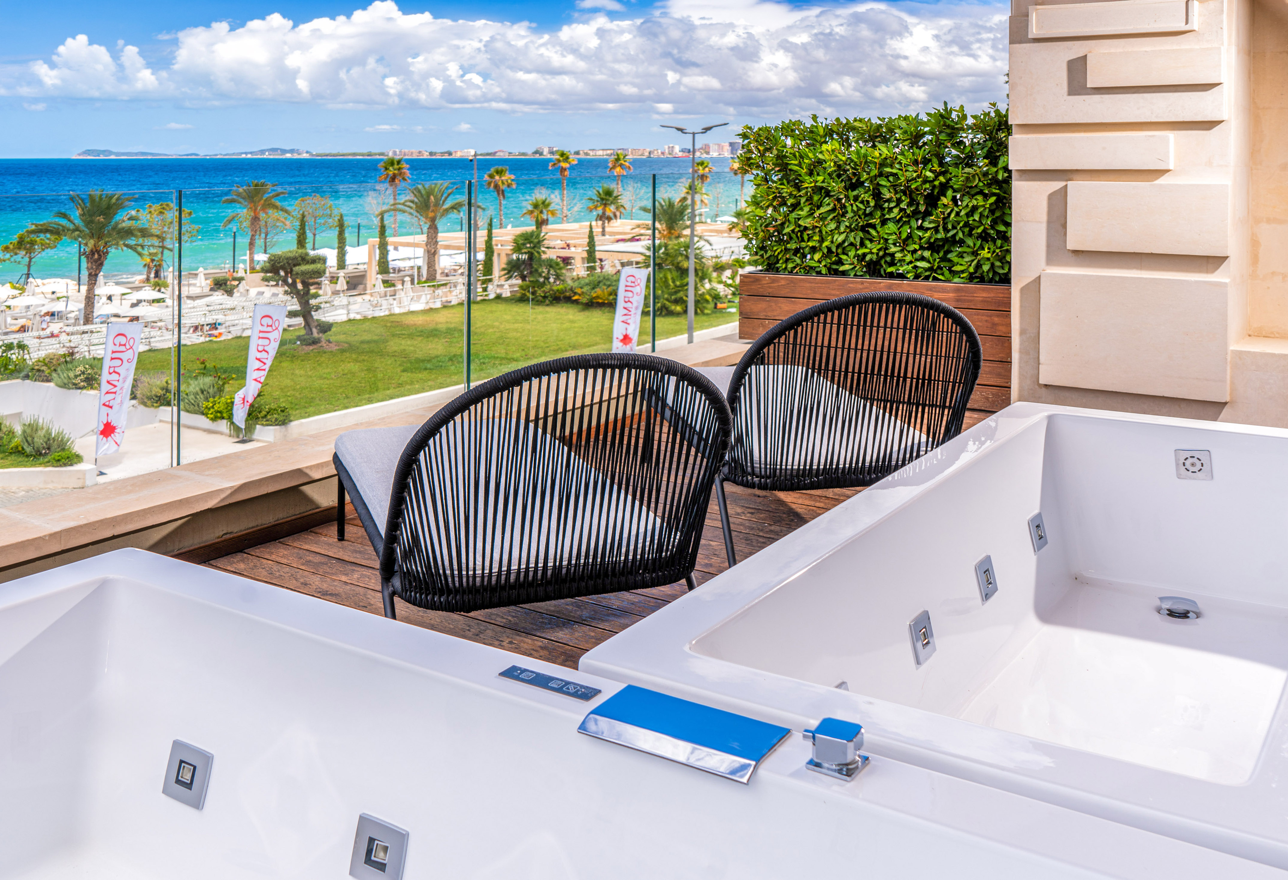 Resort balcony with two outdoor jacuzzis, chairs, beach, and blue sea view.