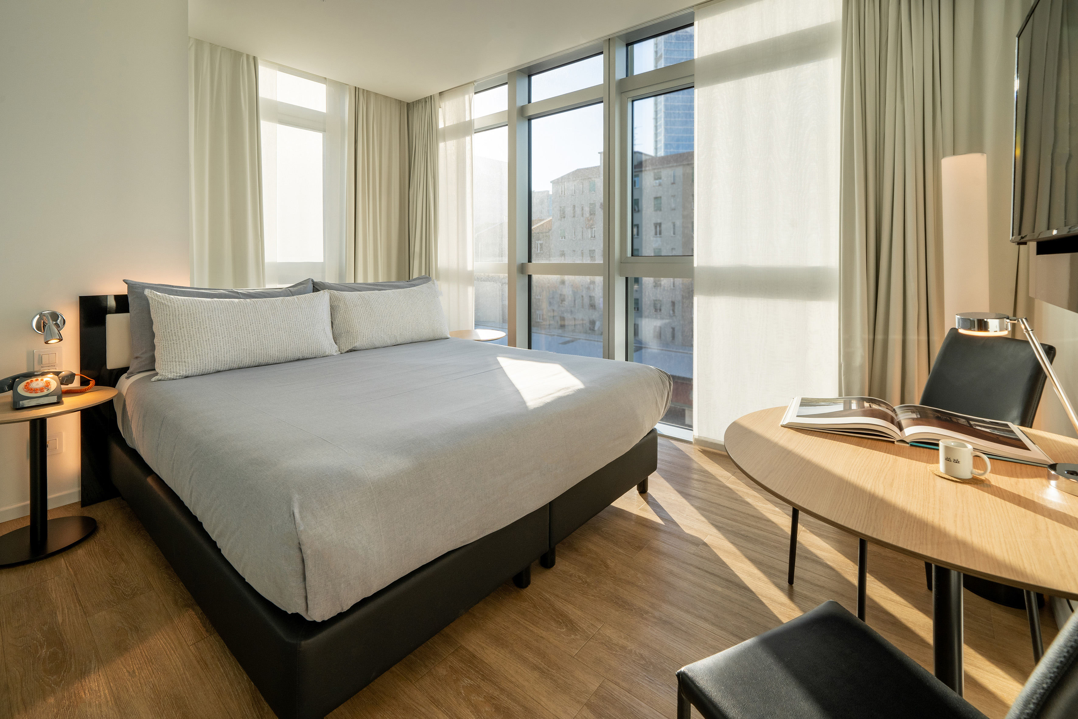 Modern hotel room with bed, large windows, city view, and natural light.