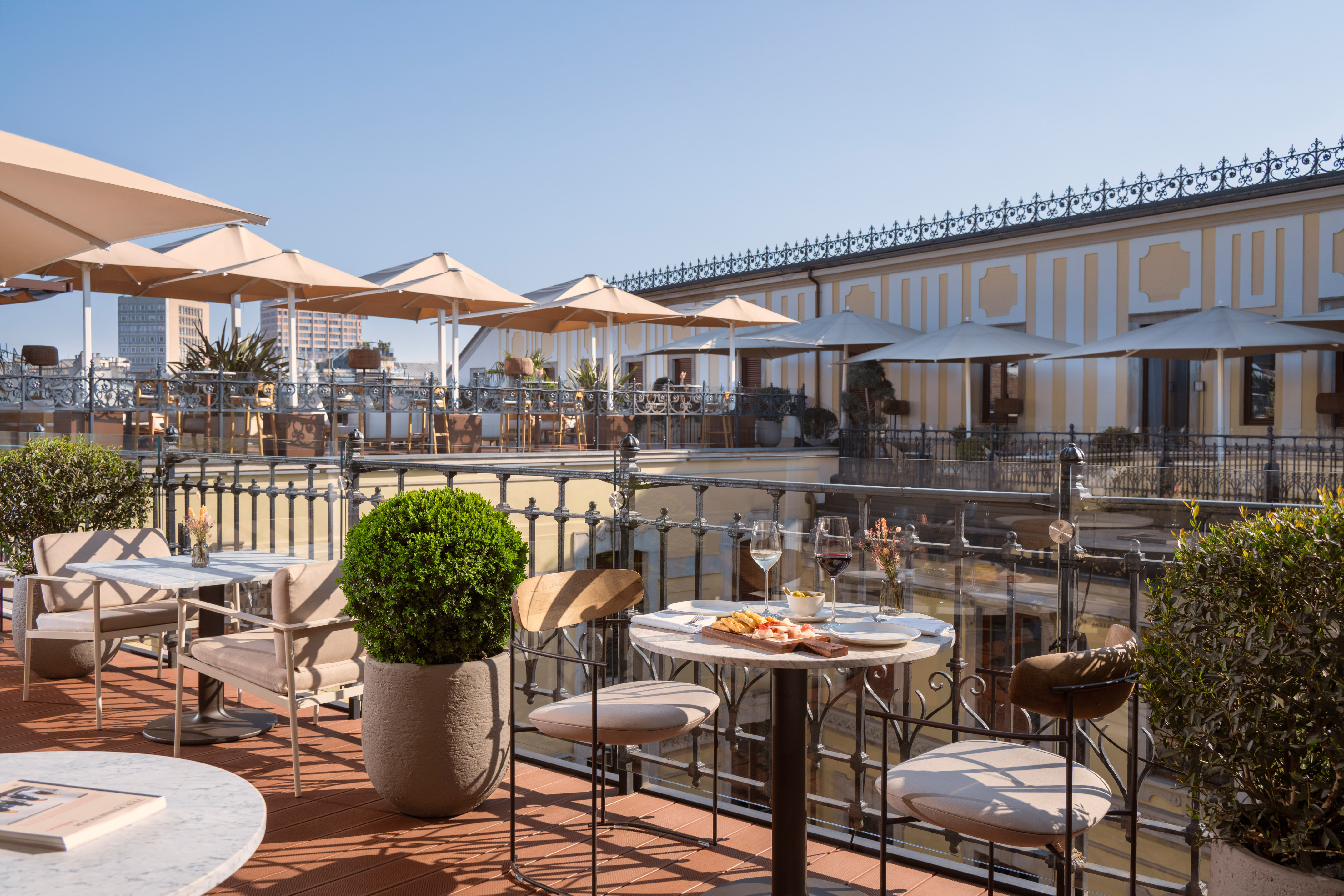 Rooftop terrace restaurant with tables, parasols, wine, food, and city view.