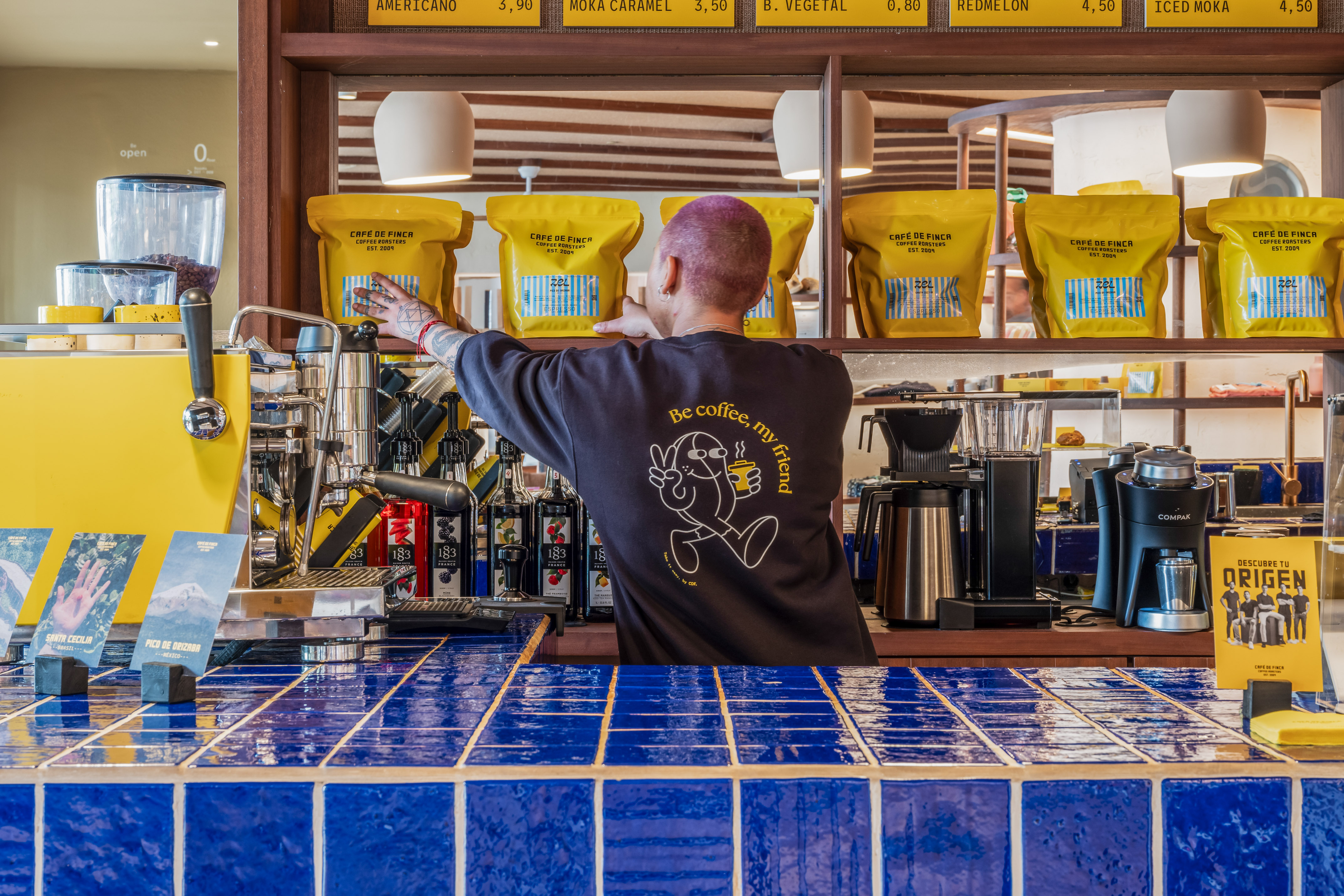 Barista arranging 'Café de Finca' coffee bags on a blue-tiled cafe counter.
