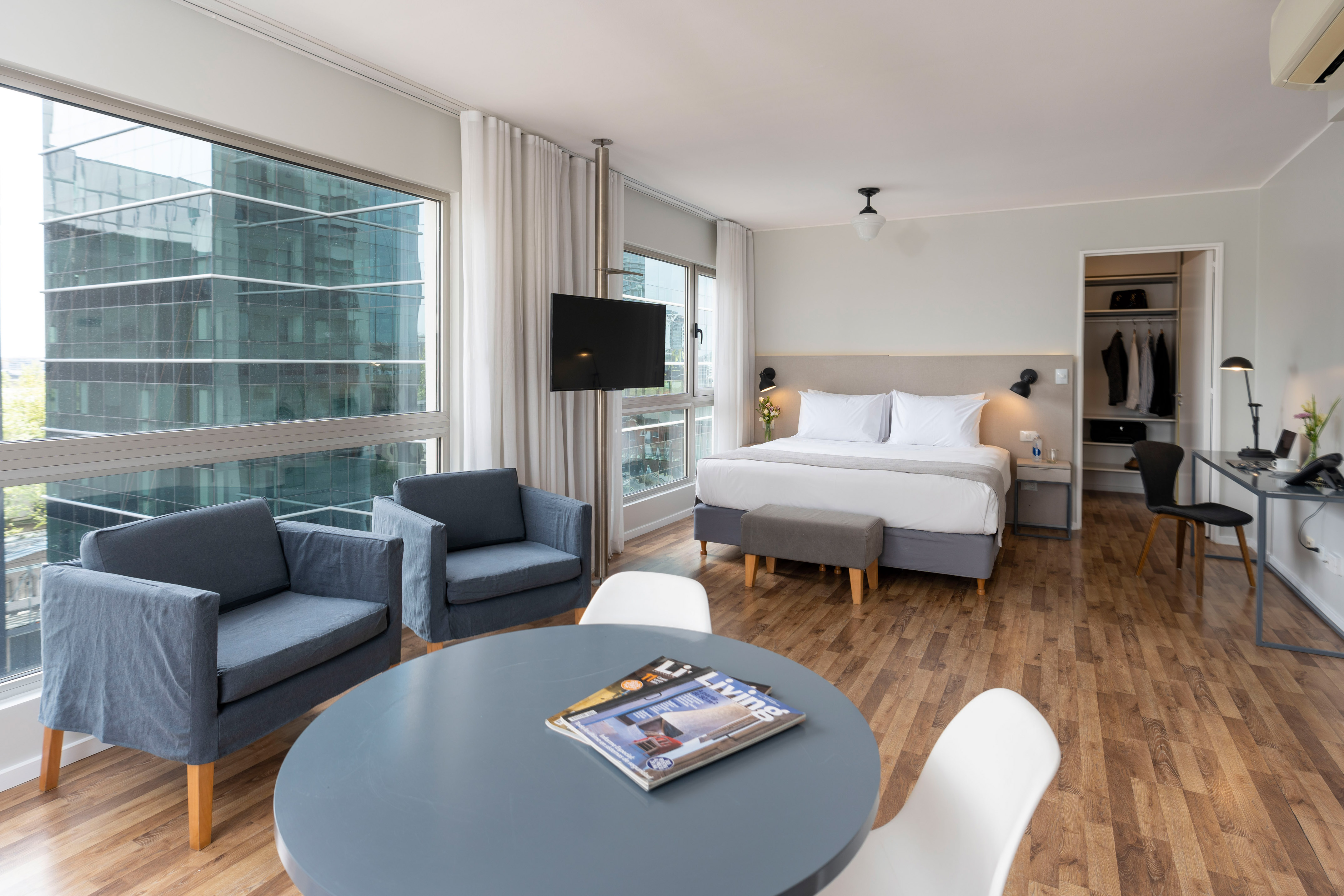 Modern hotel studio with bed, armchairs, desk, and large window city view.