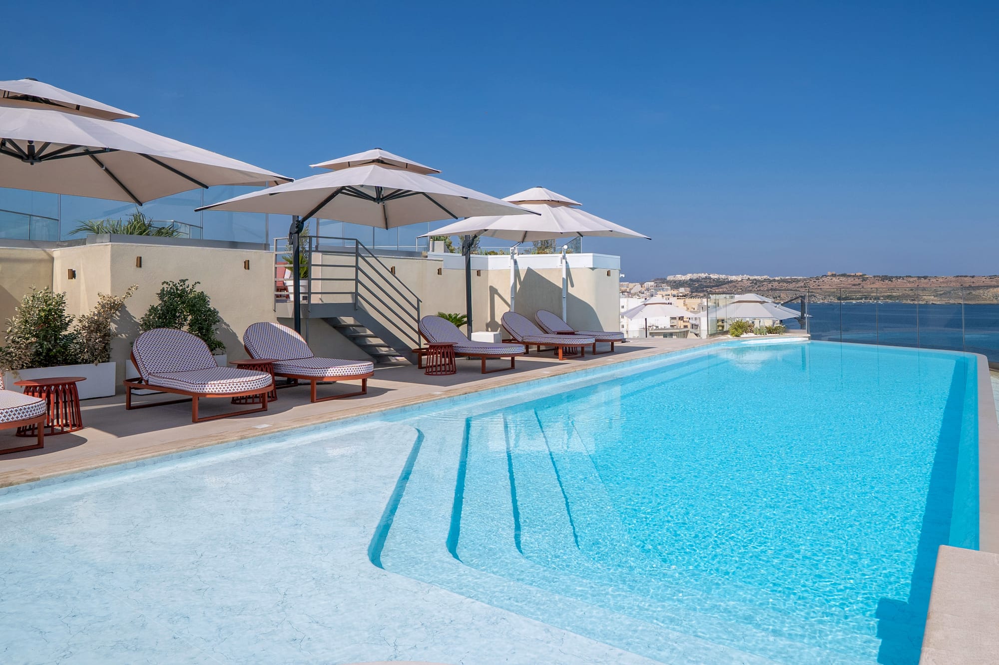 Hotel rooftop infinity pool with sun loungers, umbrellas, and sea view.