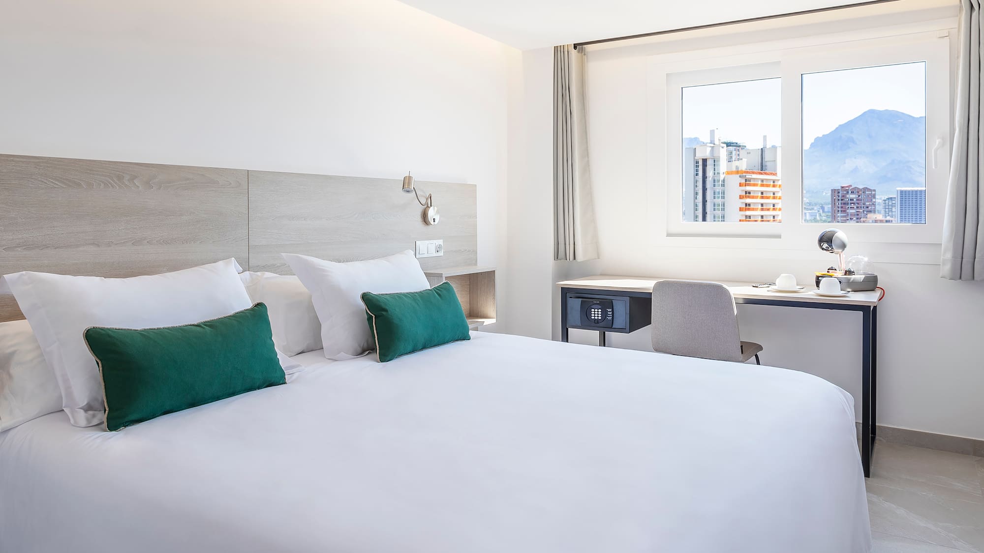 Modern hotel room with white bed, green pillows, desk, and mountain city view.