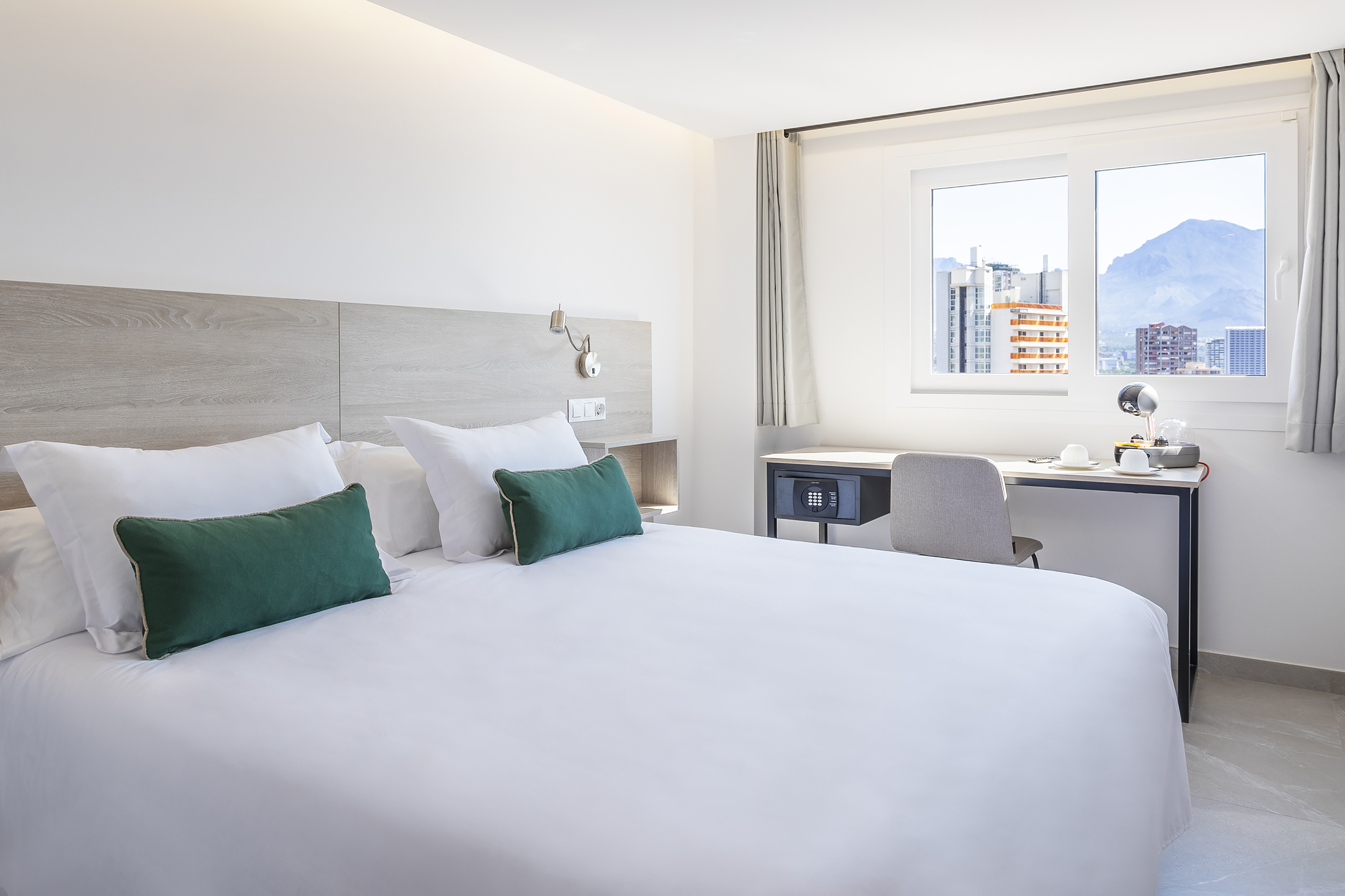 Modern hotel room with white bed, green pillows, desk, and mountain city view.