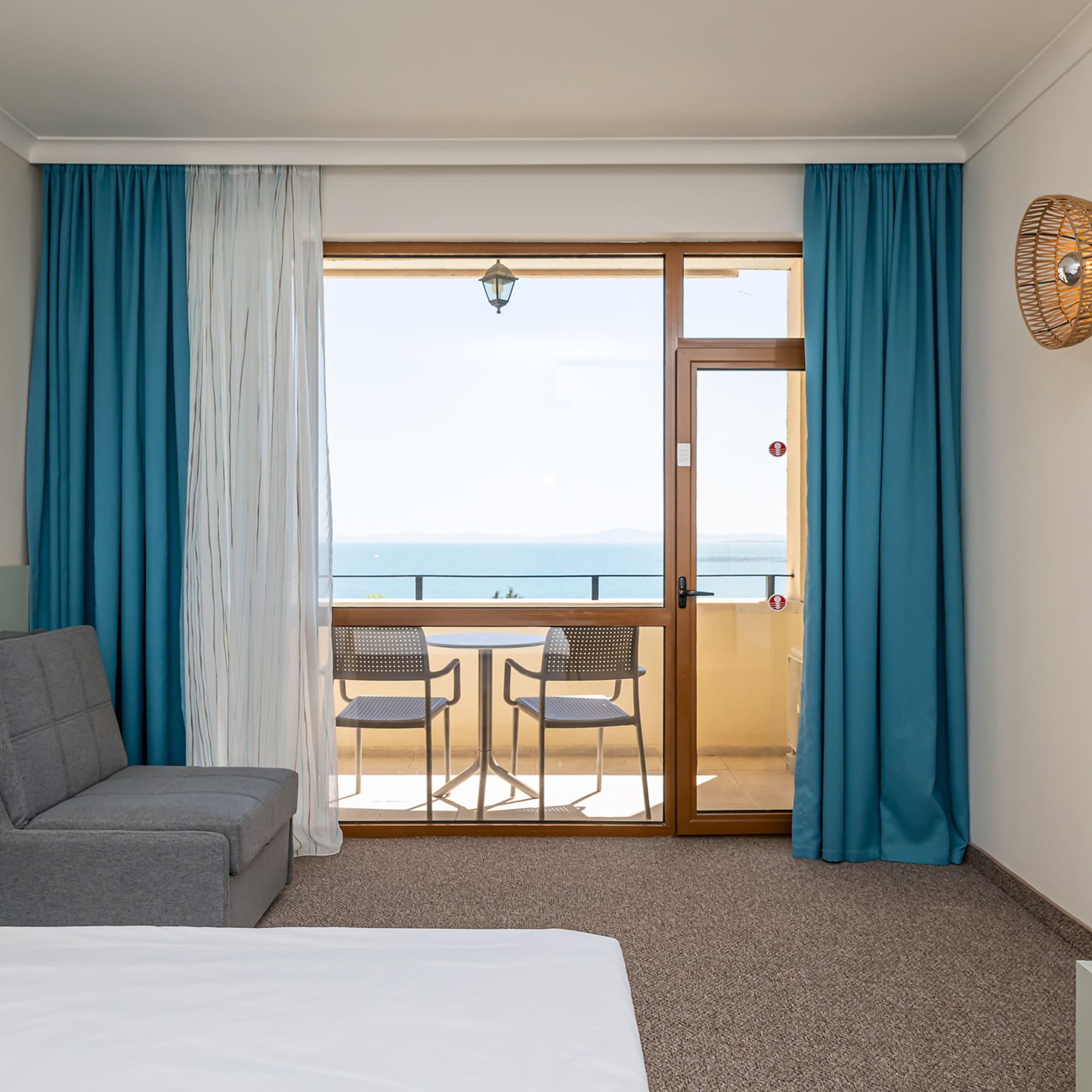 Hotel room with a sea view balcony, gray sofa bed, and blue curtains.