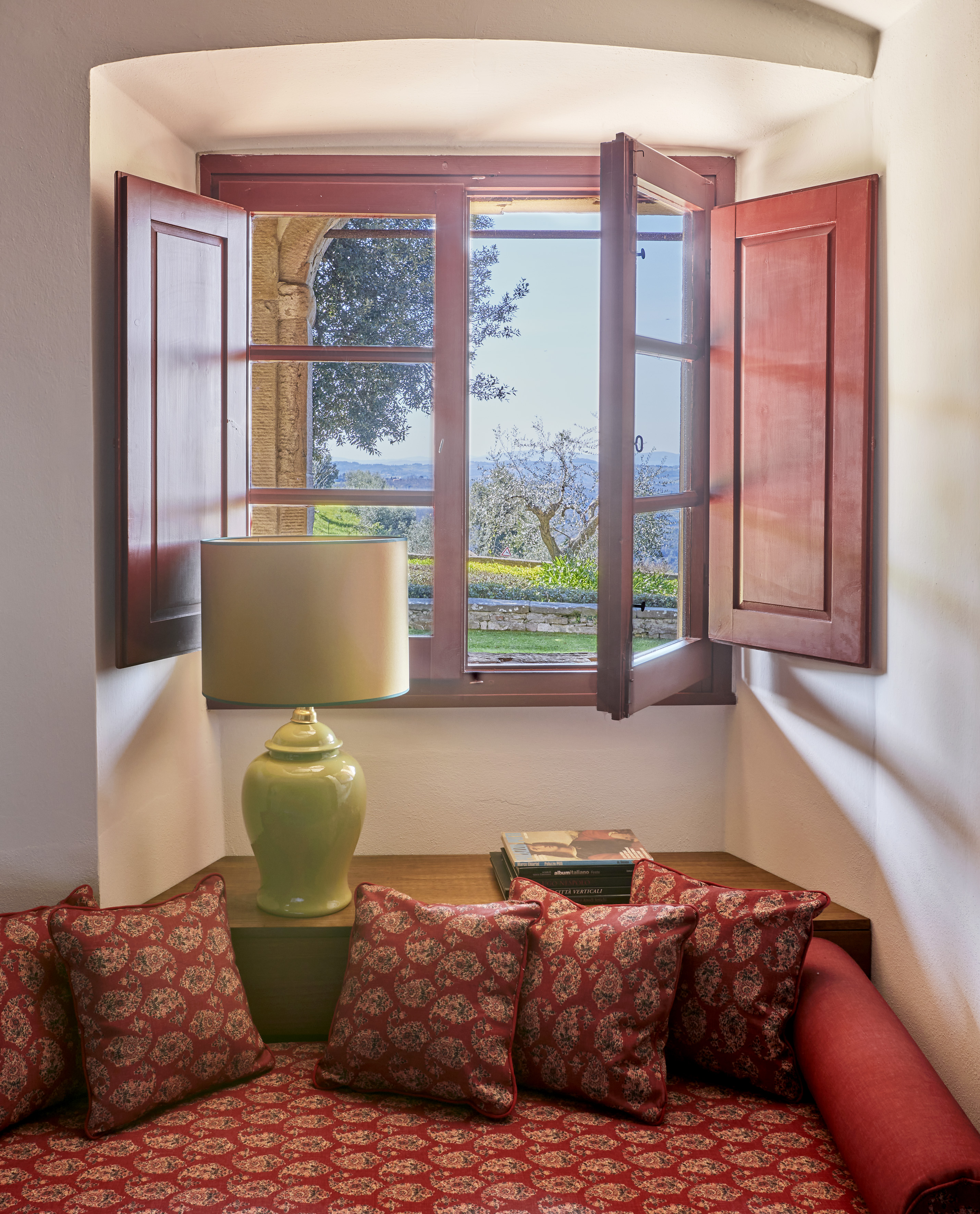 Open window with garden view, red shutters, cozy cushions, and lamp.
