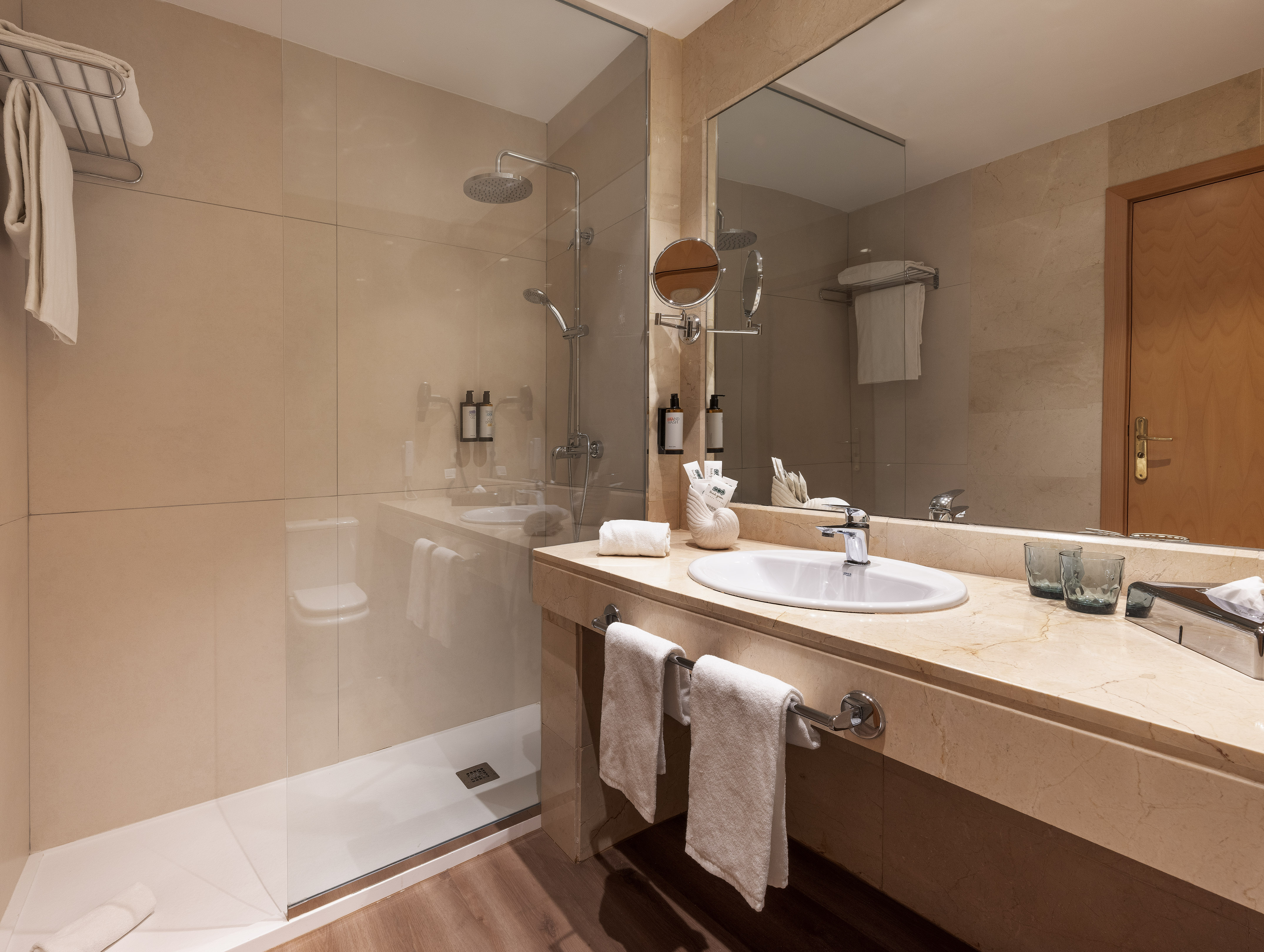 Modern hotel bathroom with glass shower, beige tiles, and marble vanity.