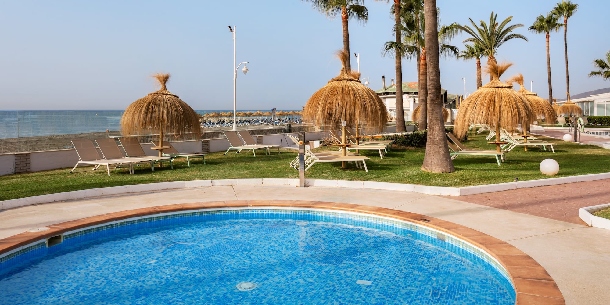 Sunny resort pool with beach, palm trees, and straw umbrellas.