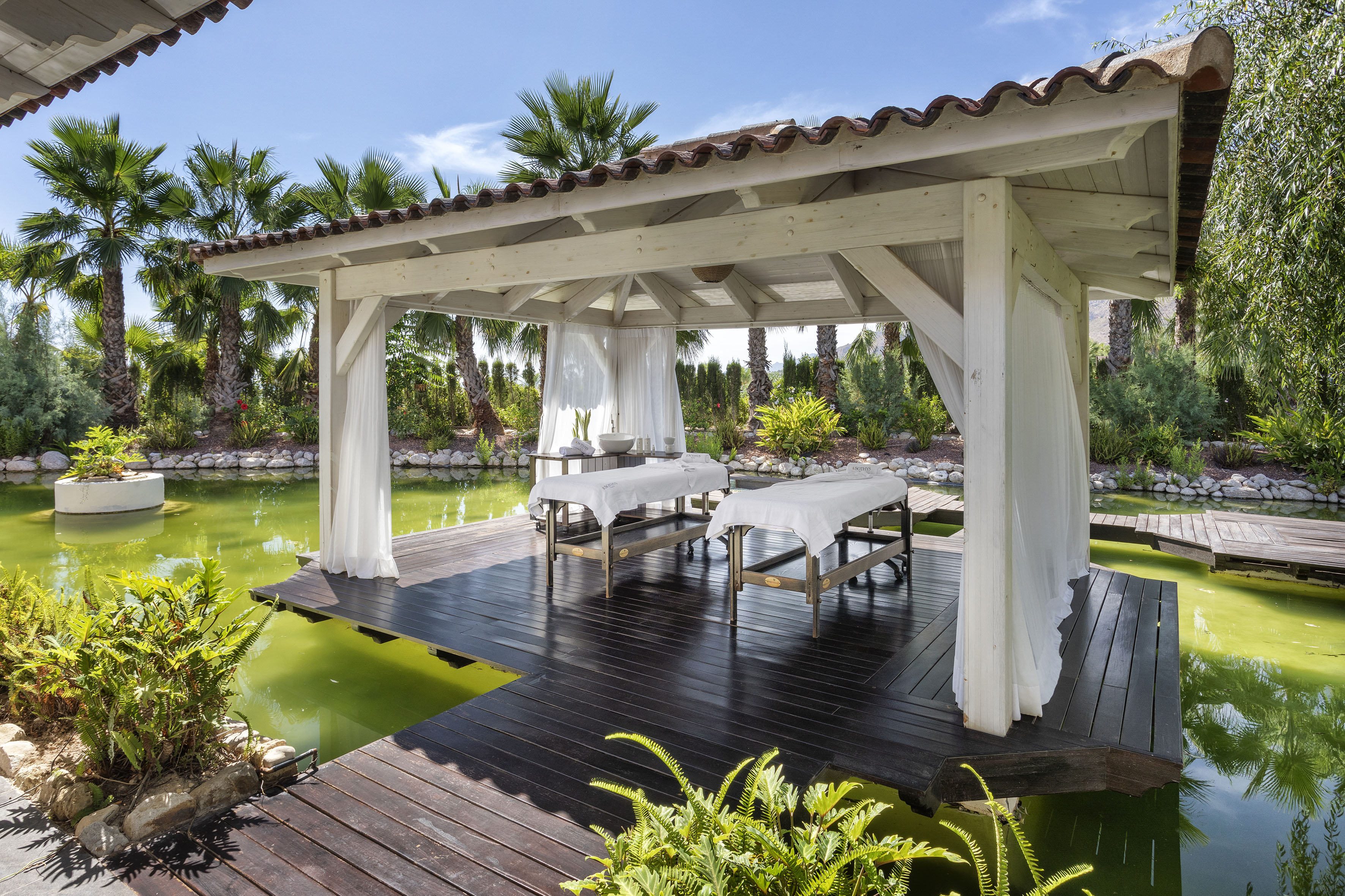 Outdoor spa gazebo with two massage tables over green pond, palm trees.