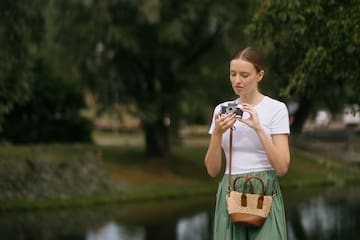 Woman holding vintage camera outdoors. White shirt, green skirt, by river.