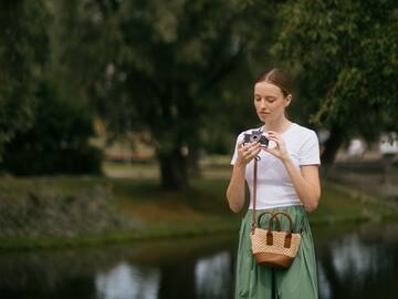 Woman holding vintage camera outdoors. White shirt, green skirt, by river.