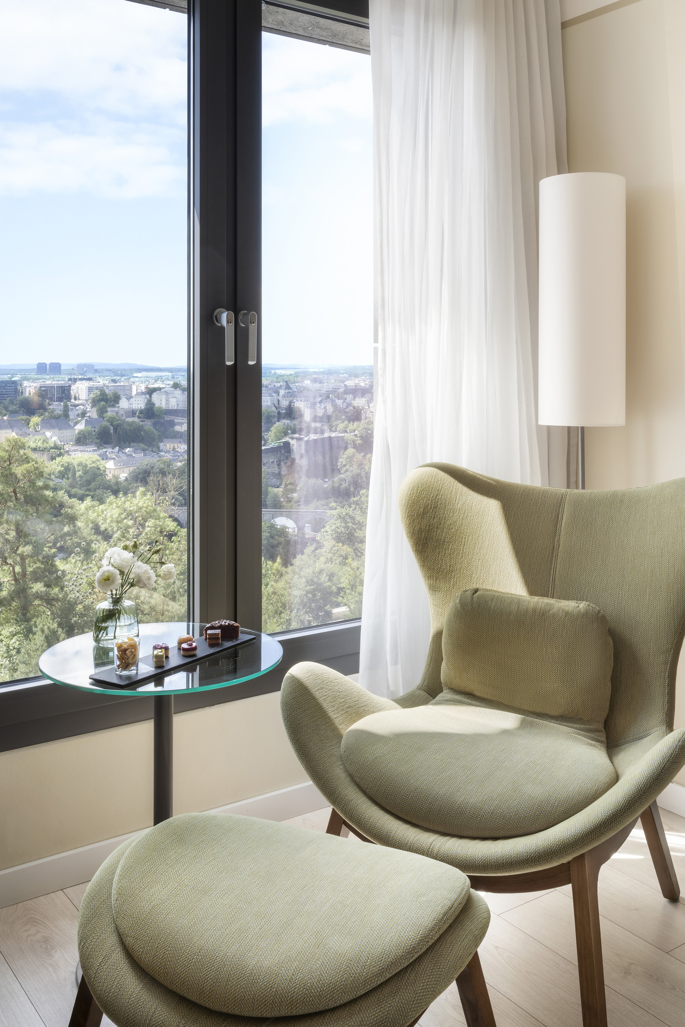 Green armchair, ottoman by window with city view and table of desserts.