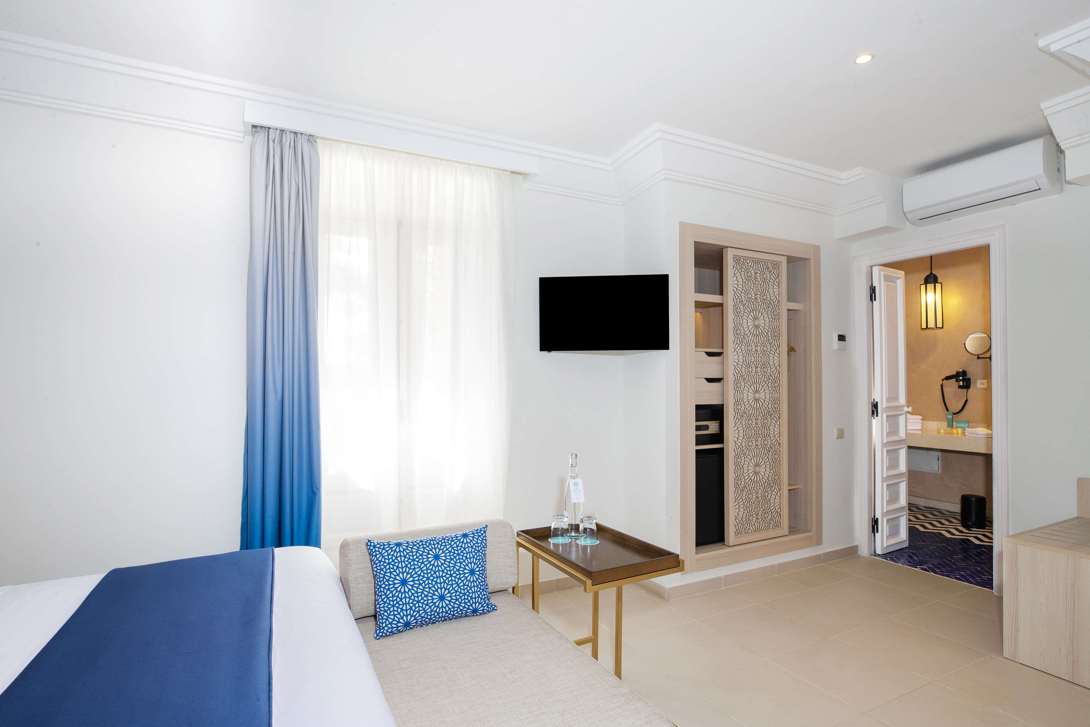 Bright hotel room with blue decor, TV, patterned closet, and open bathroom. - Sol Room cu balcon - SOL Oasis Marrakech, Marrakech