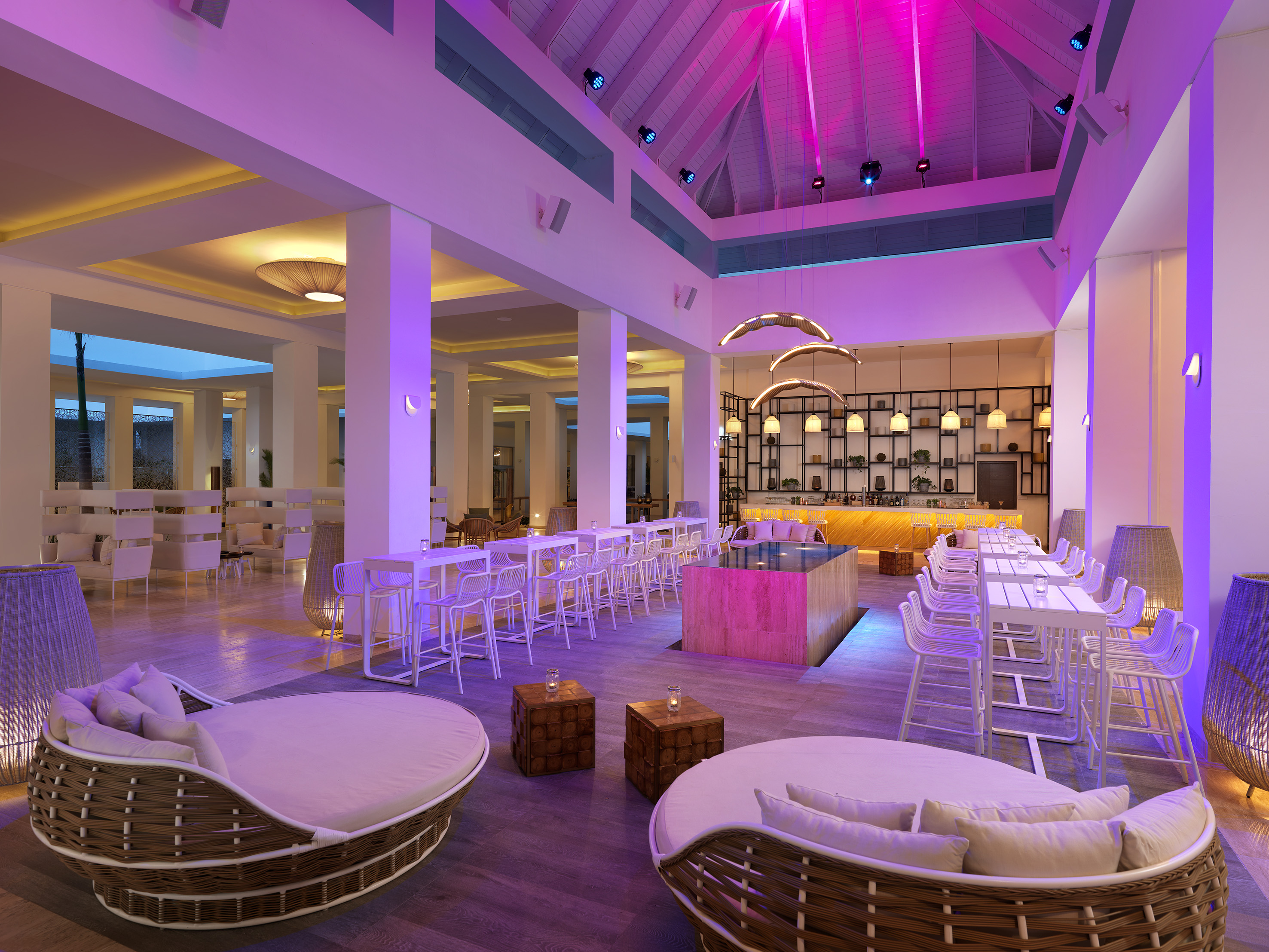 Vibrant lounge bar with pink lighting, modern high tables, and wicker daybeds.
