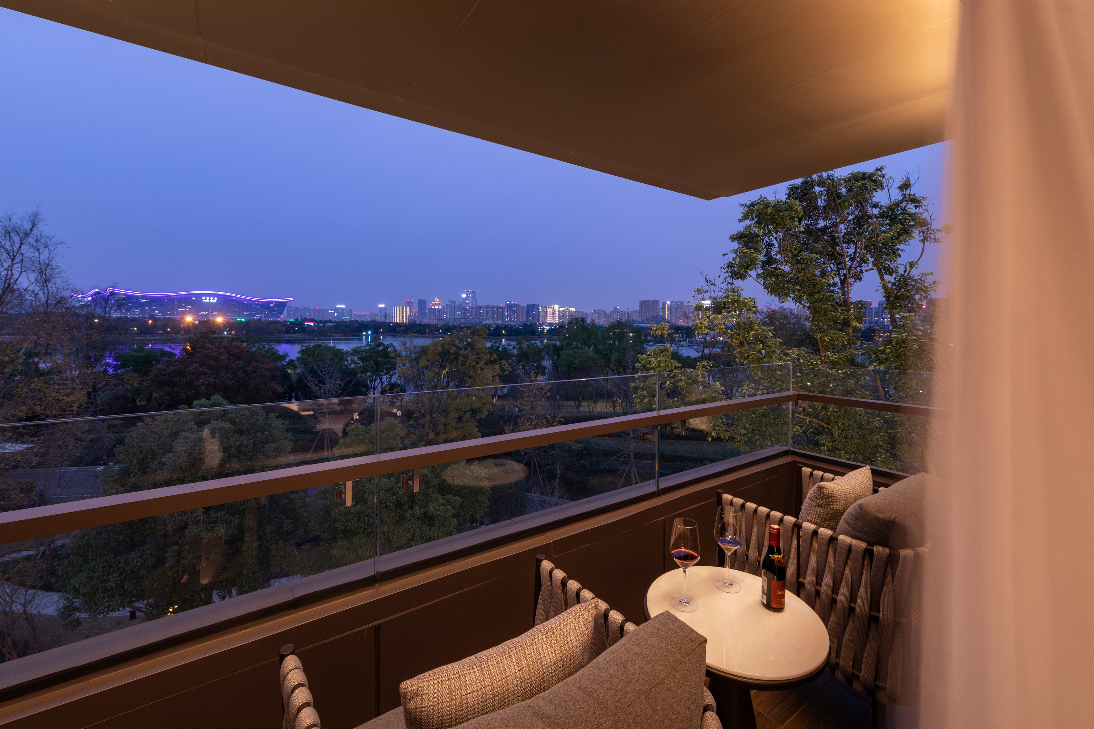 Balcony at dusk overlooking a city skyline, lake, with wine and chairs.