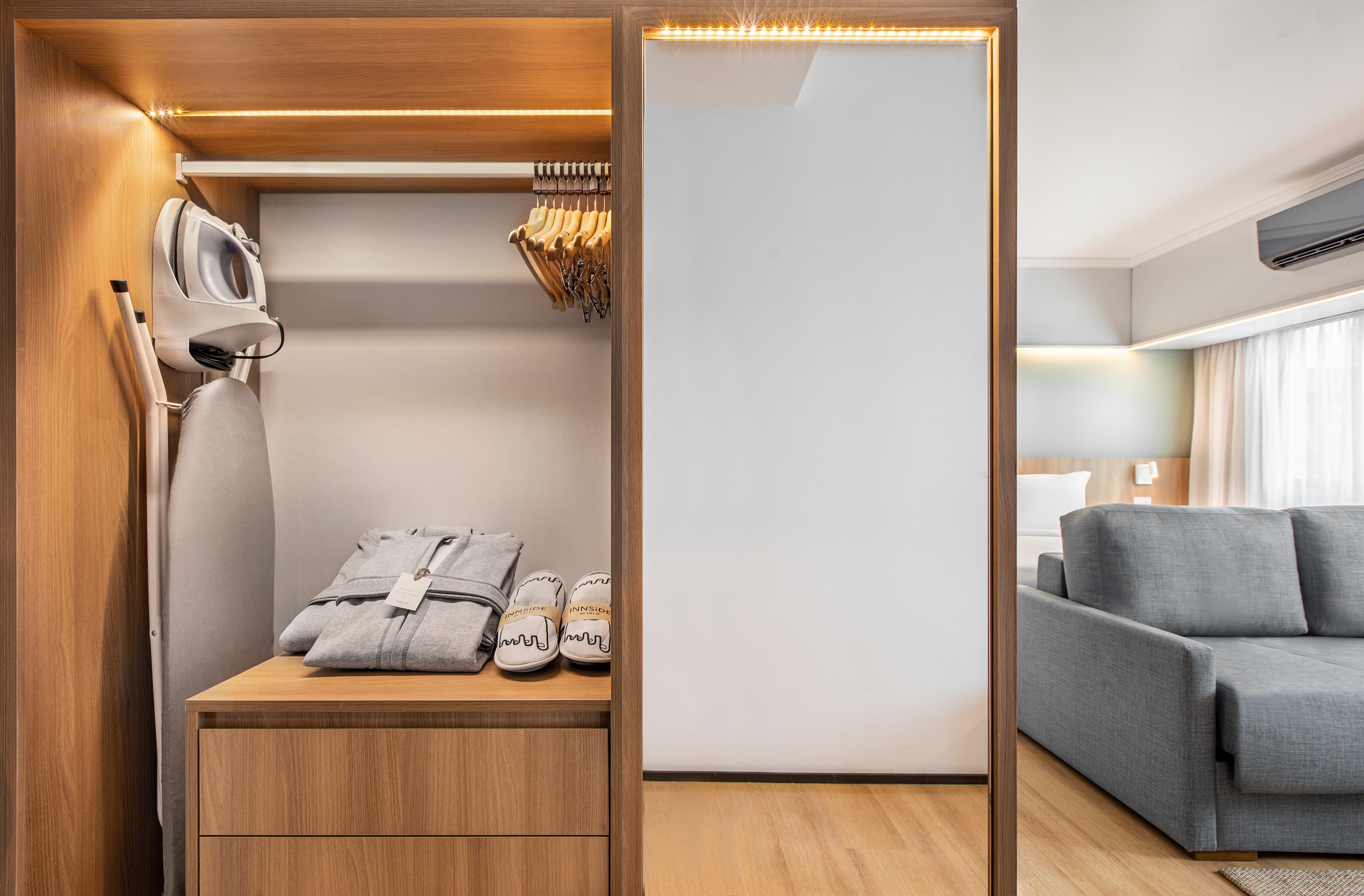 Hotel closet with iron, ironing board, robes, slippers, and modern room view.