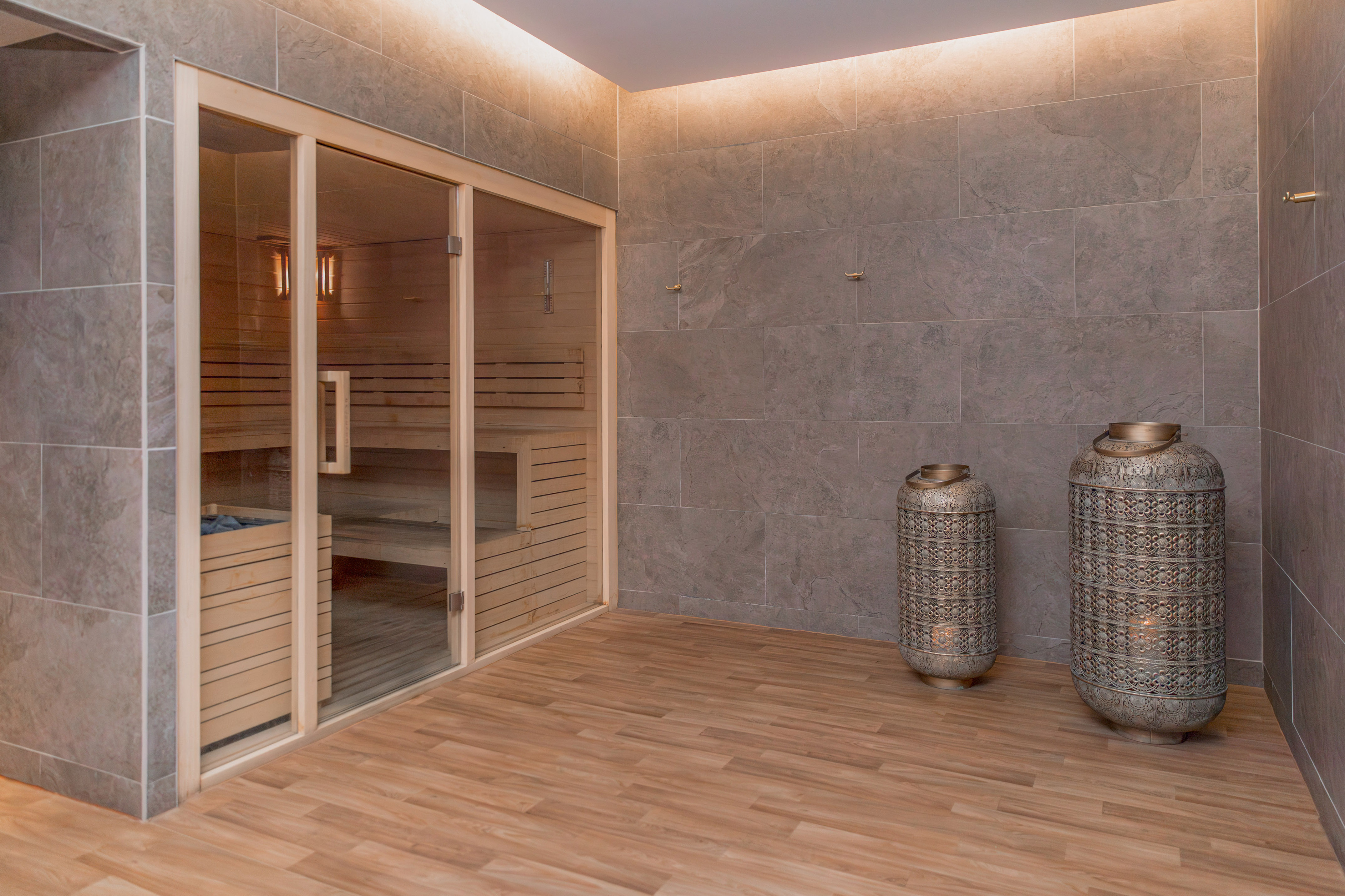 Modern wood and glass sauna, grey tiled walls, wooden floor, and lanterns.
