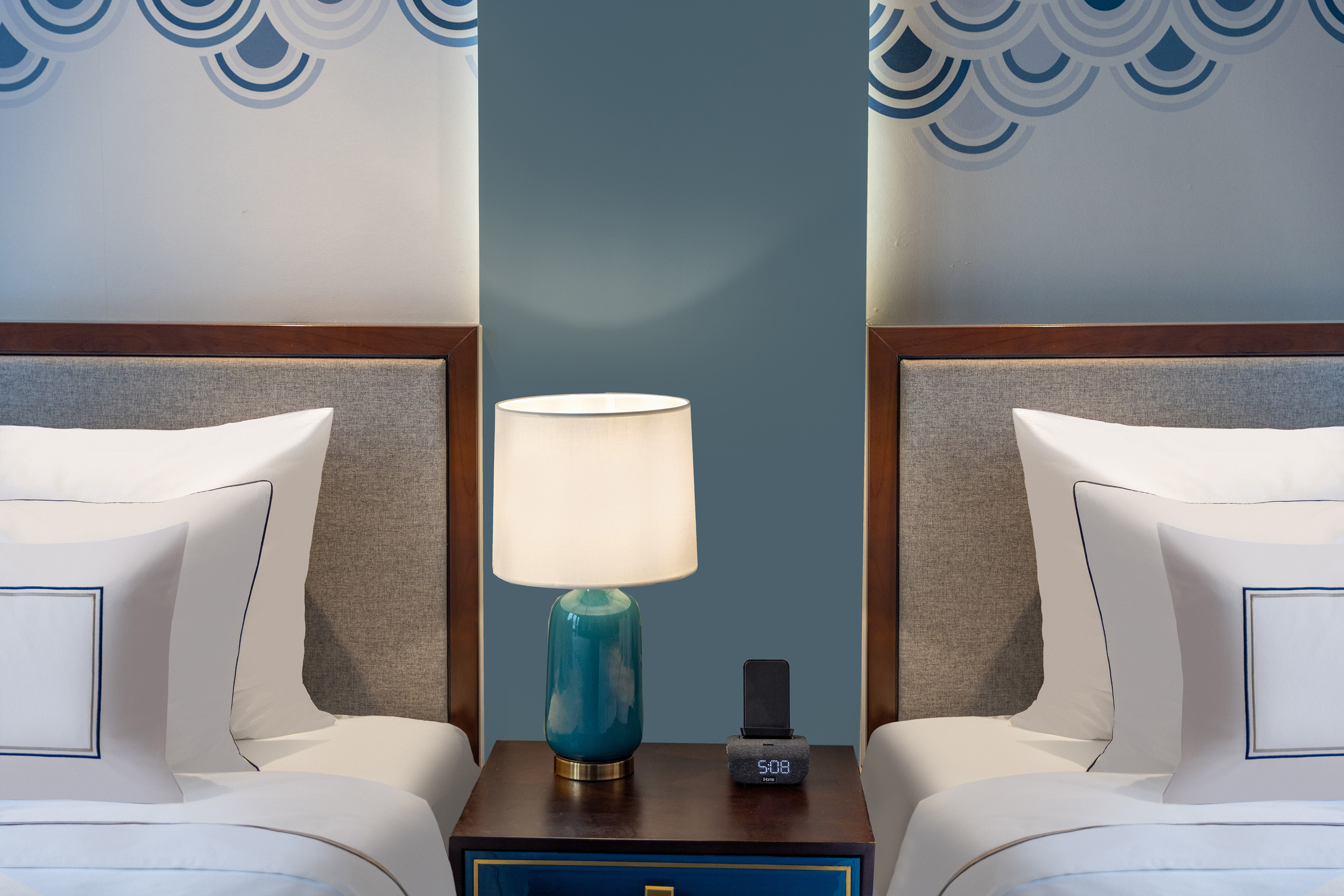 Hotel bedroom with twin beds, nightstand, teal lamp, blue wave decor.
