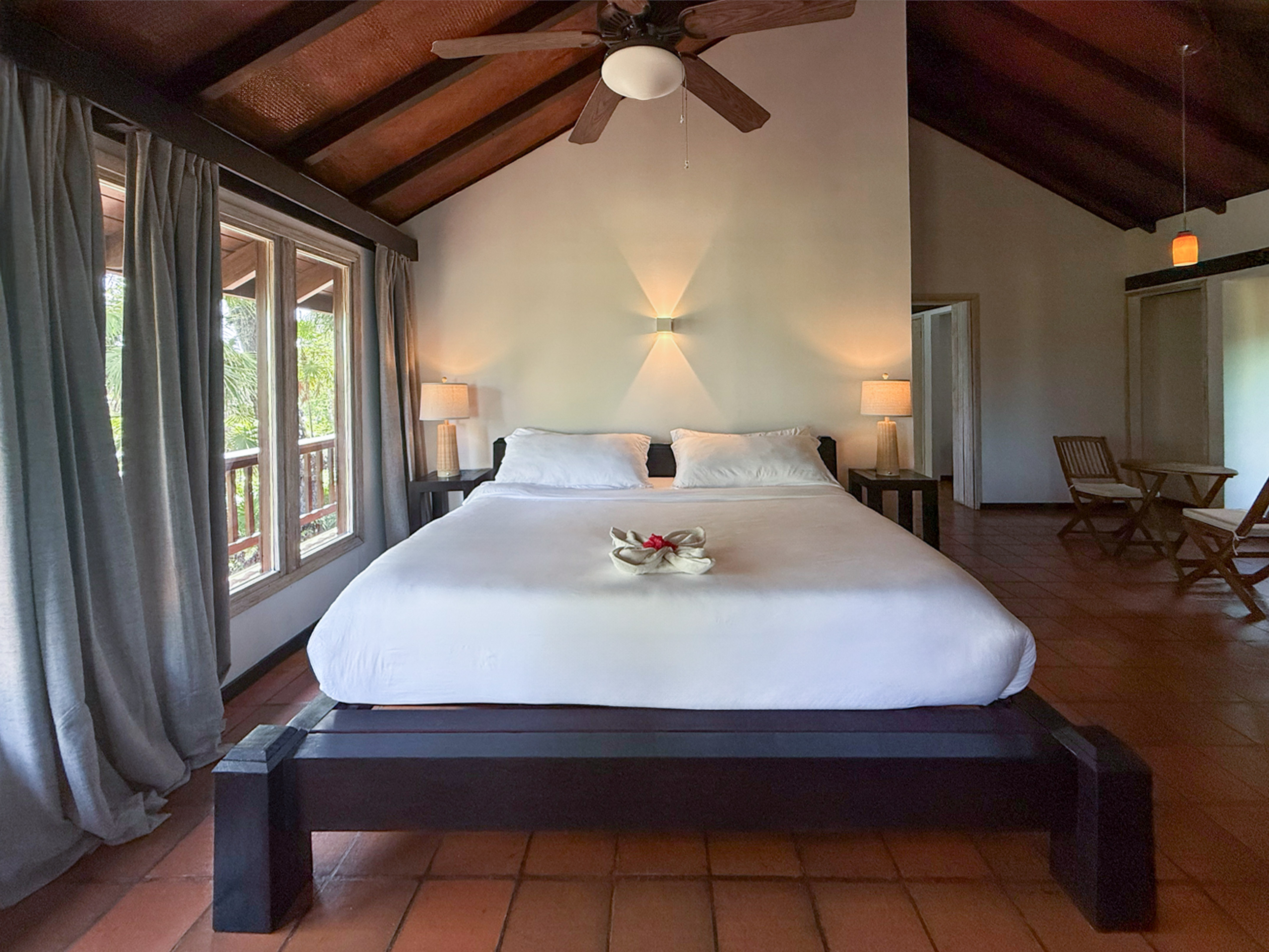 Rustic tropical bedroom with vaulted wood ceiling, queen bed, and balcony view.