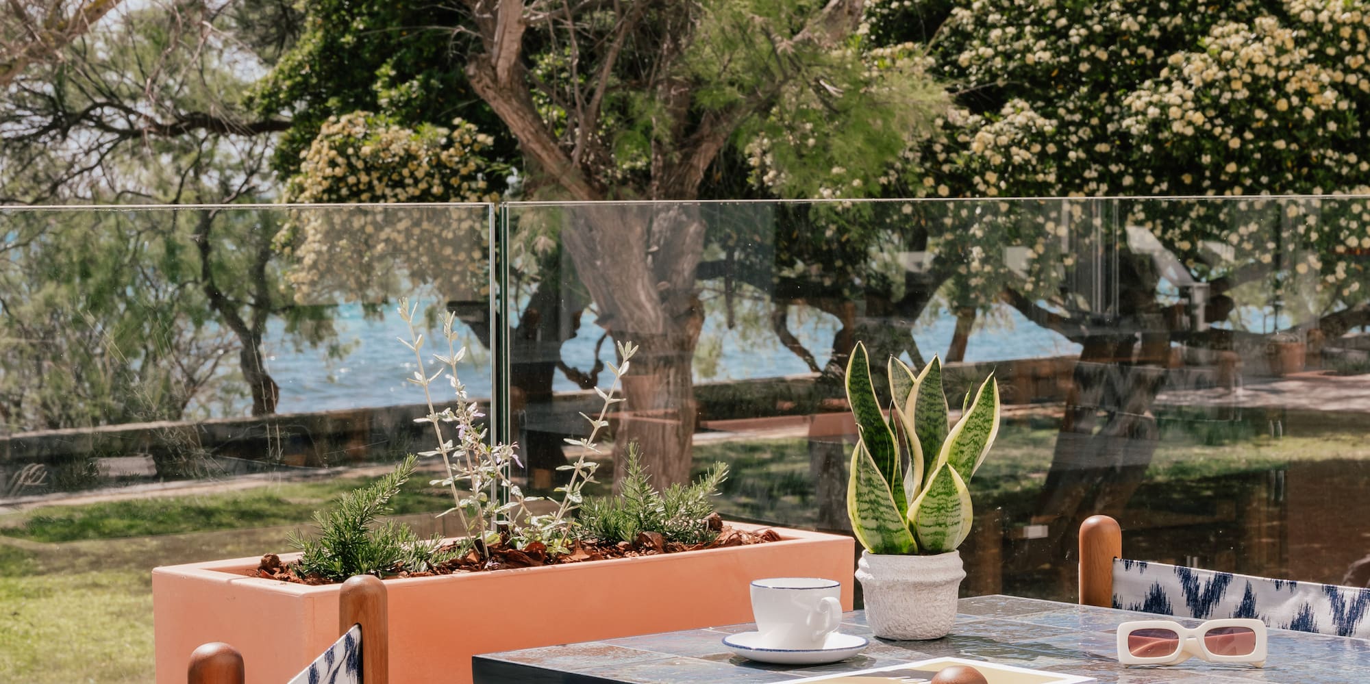 Resort patio dining area with sea view, blue table, wooden chairs, and plants.