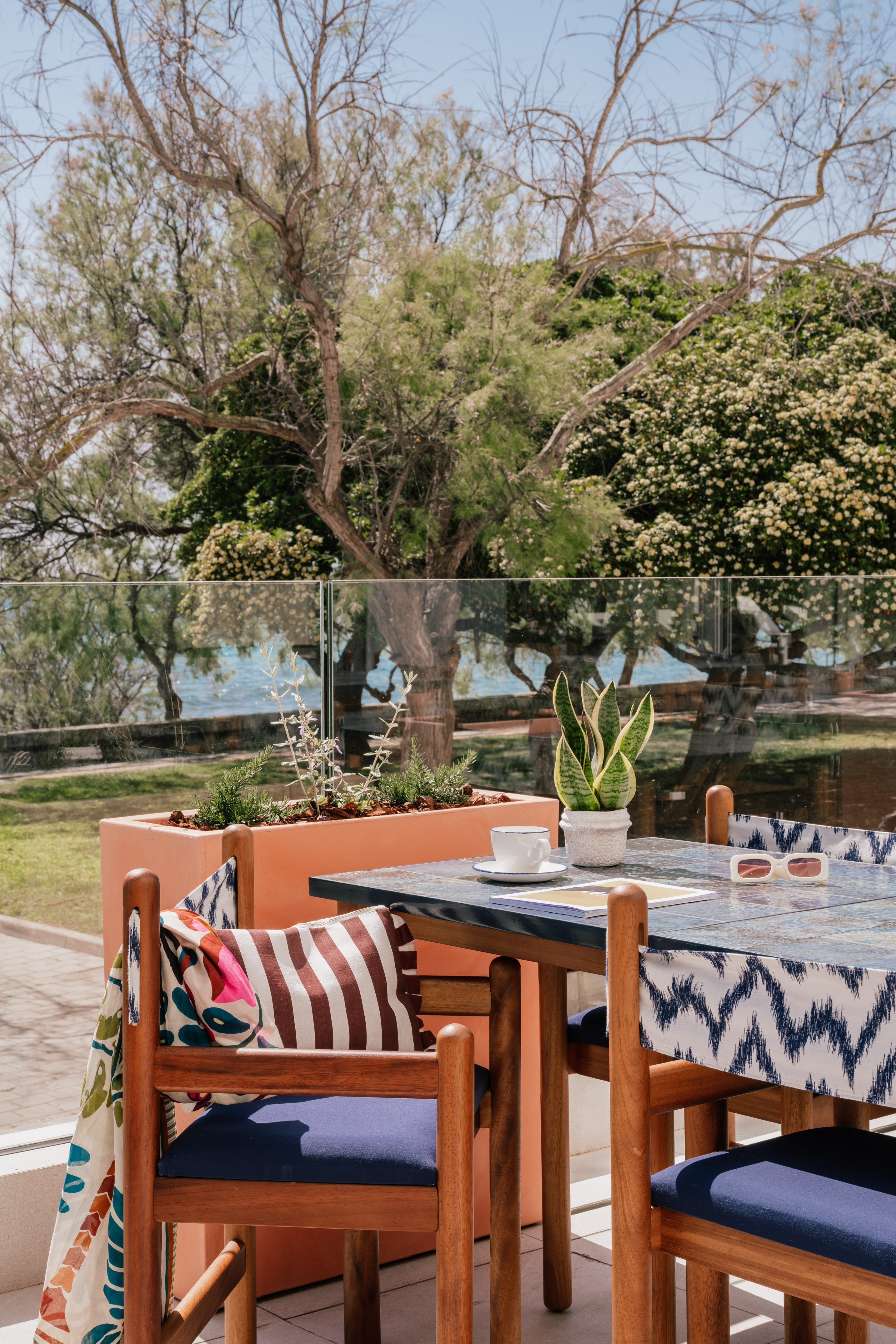Resort patio dining area with sea view, blue table, wooden chairs, and plants.