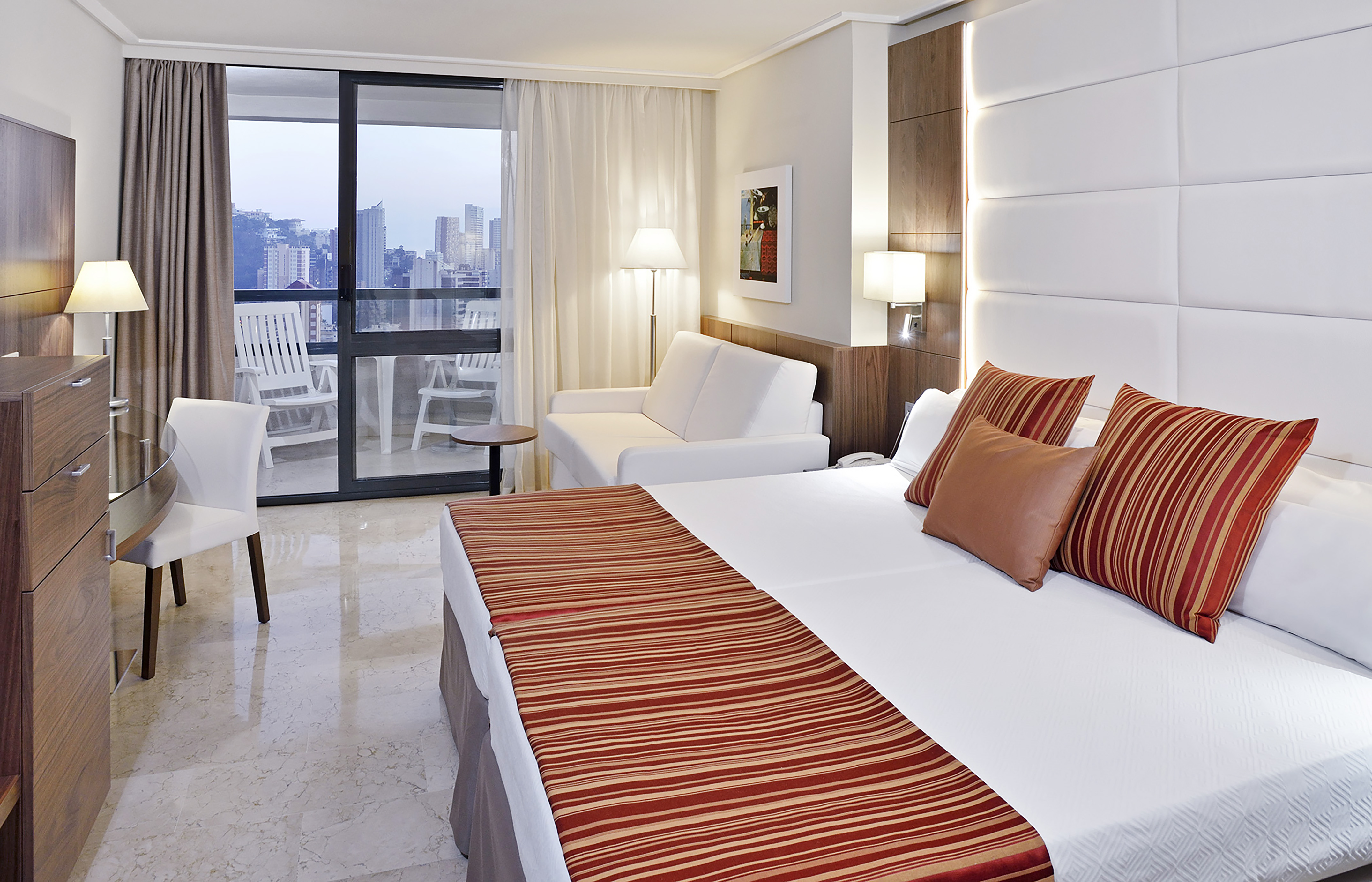 Modern hotel room with bed, sofa, desk, and city view balcony.