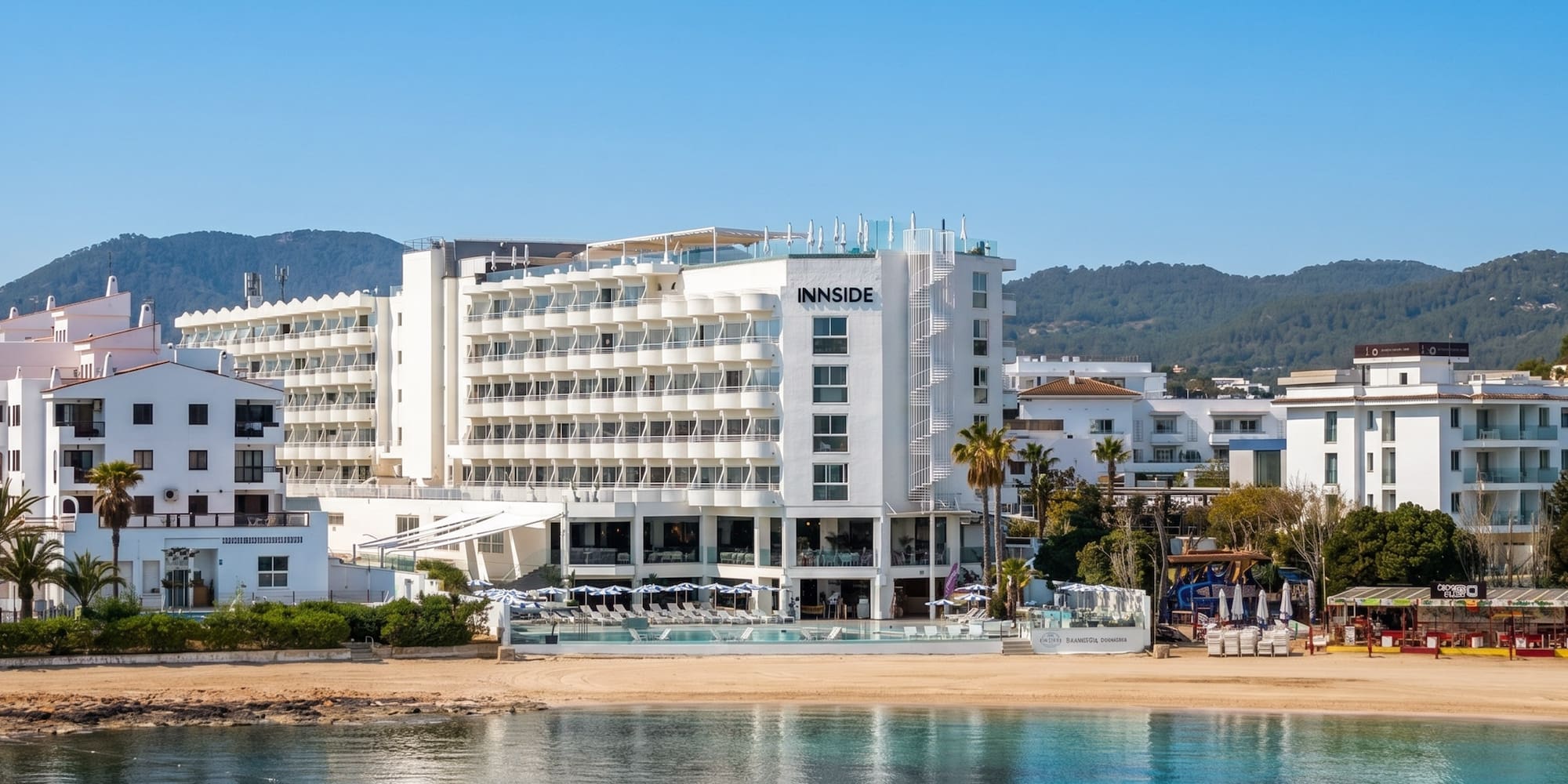 INNSIDE beach hotel on a sunny coast with turquoise water and green hills.