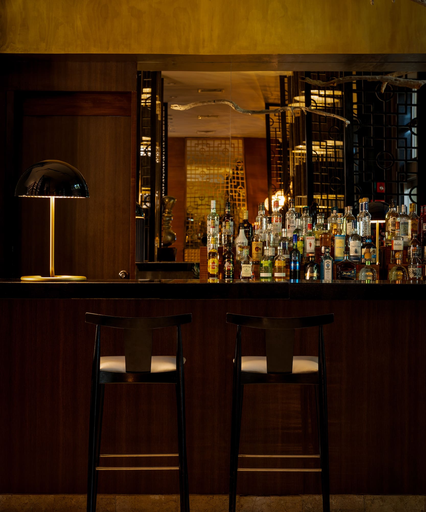Elegant hotel bar with liquor bottles, two bar stools, and warm lamp.