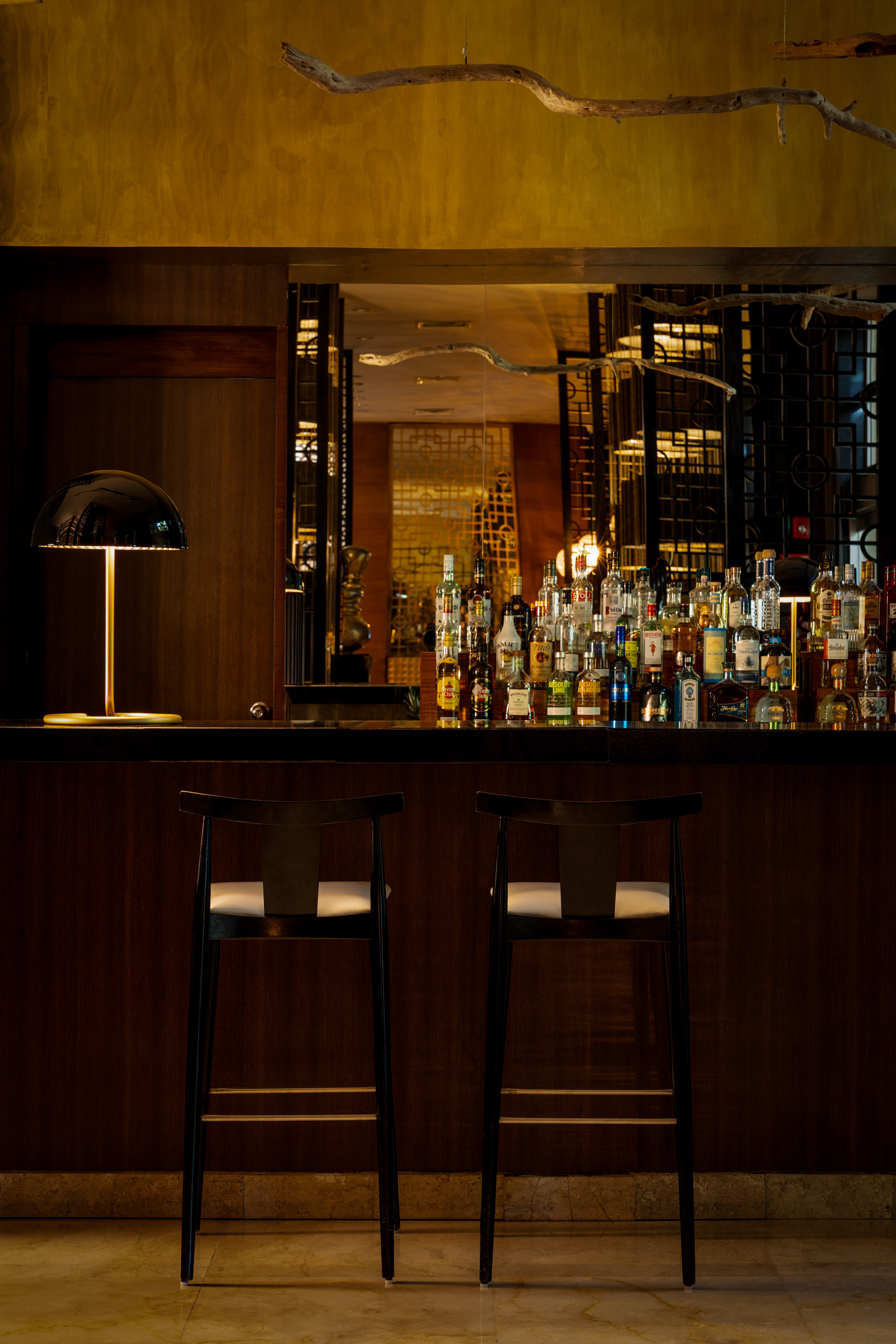 Elegant hotel bar with liquor bottles, two bar stools, and warm lamp.