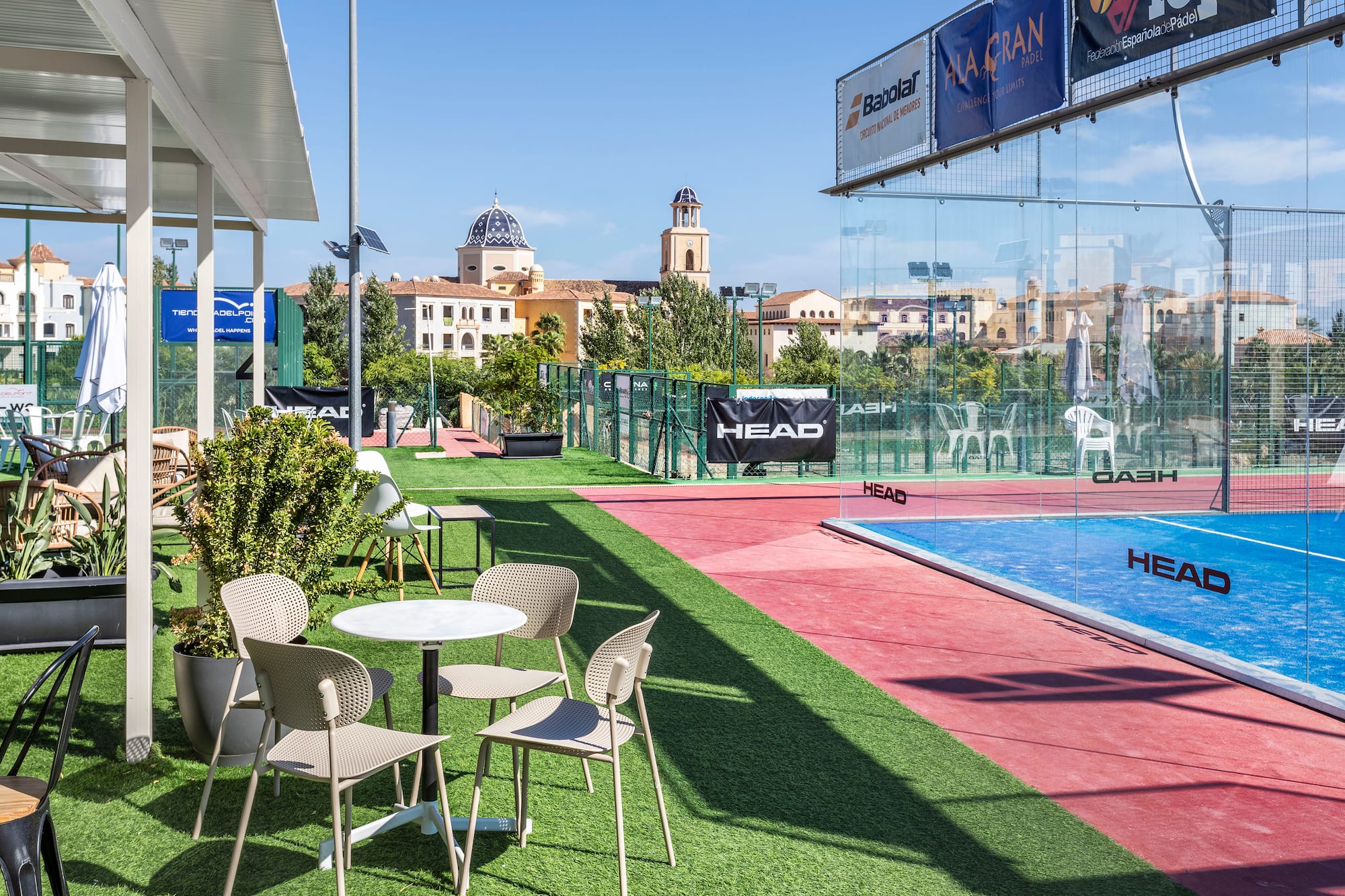 Outdoor padel court with cafe area on grass, classic Mediterranean buildings.