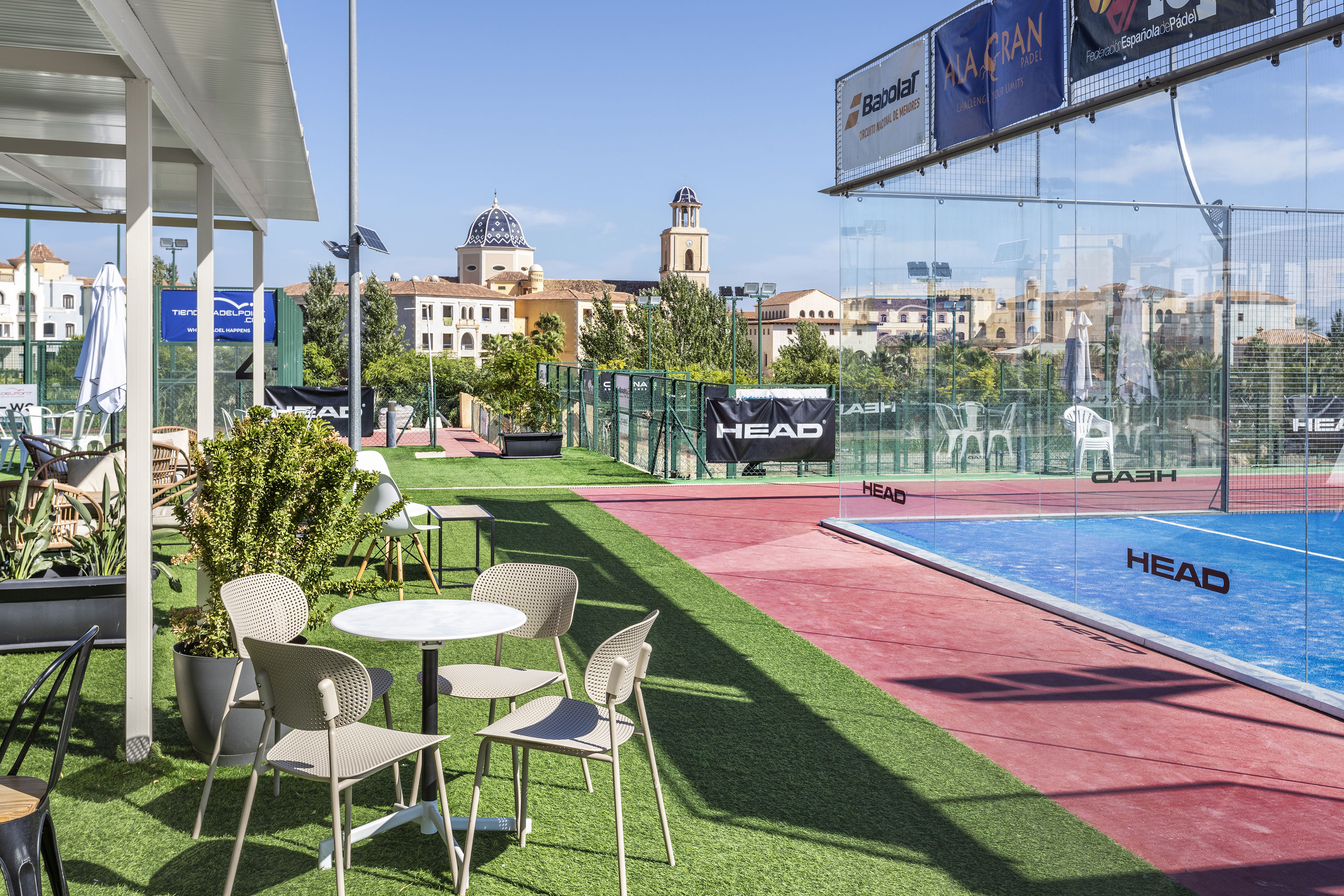 Outdoor padel court with cafe area on grass, classic Mediterranean buildings.