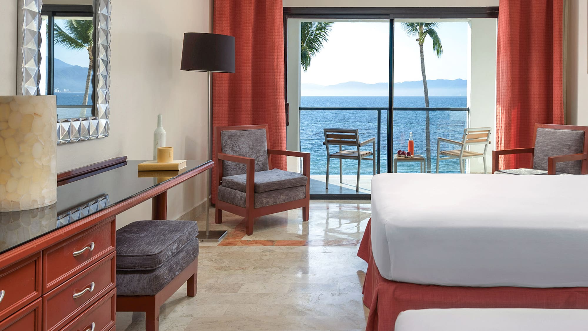 a room with a bed and chairs and a table with a view of the ocean