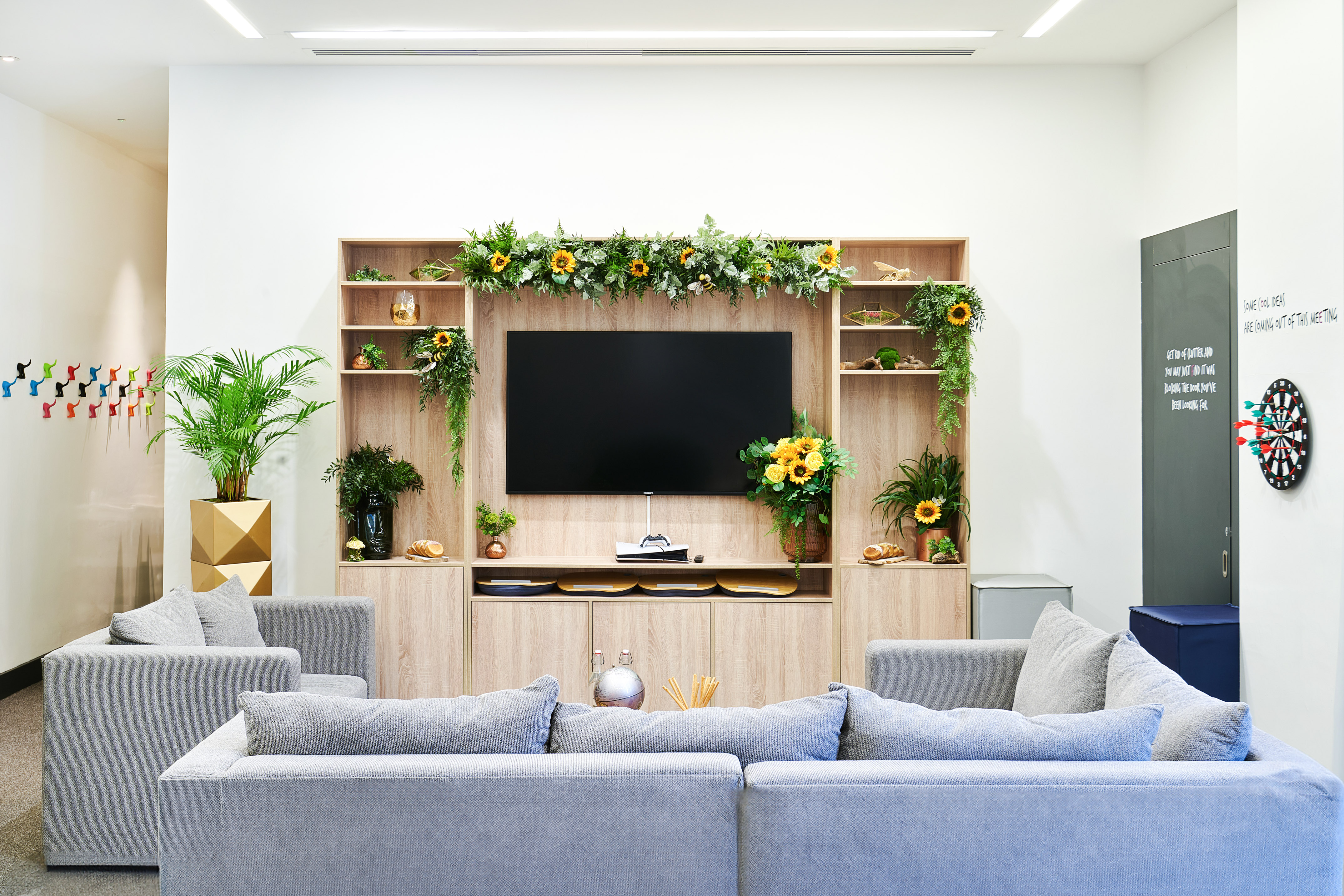 Vibrant office break room with sofas, TV, plants, sunflowers, and dartboard.
