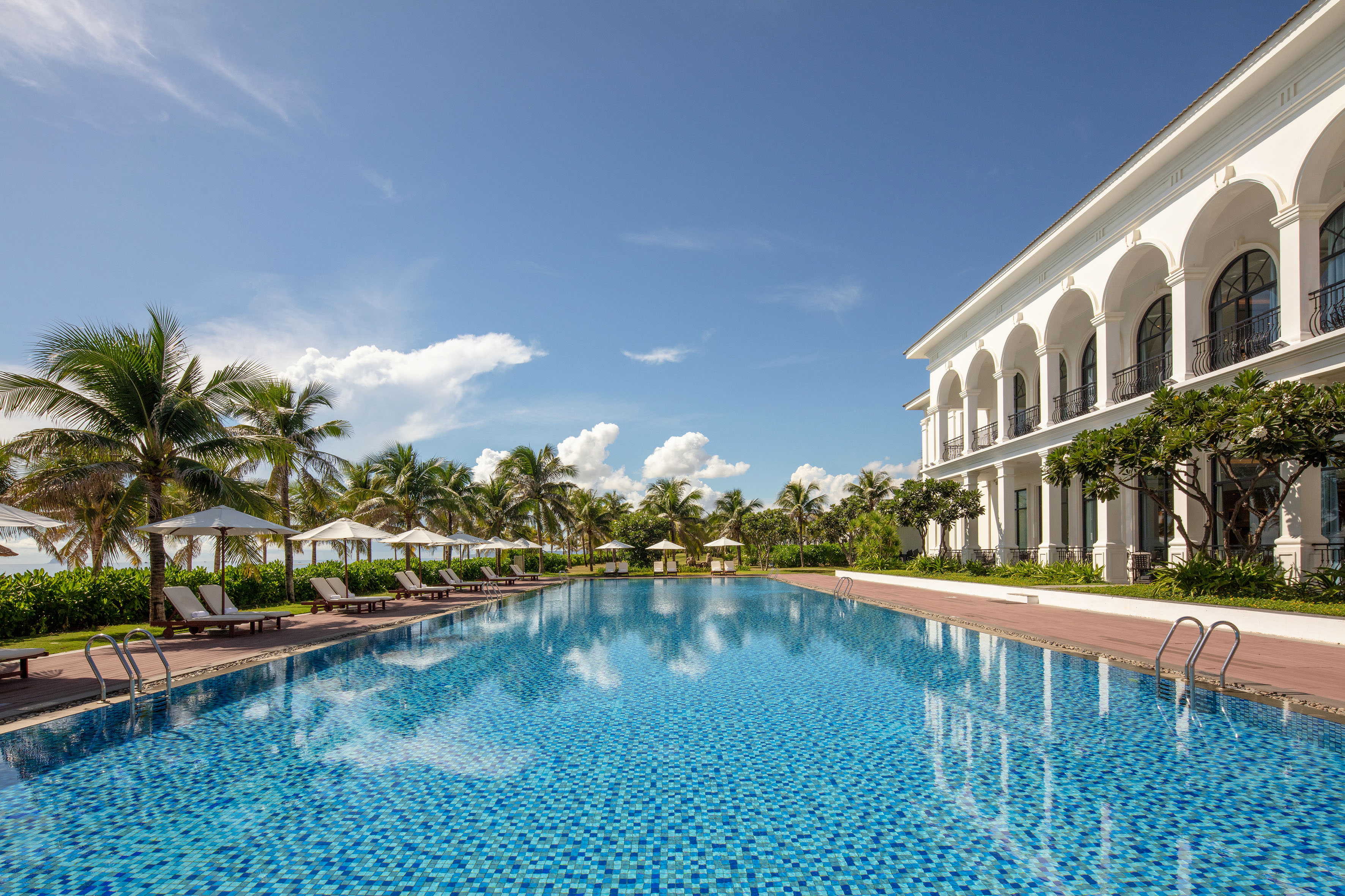 Tropical resort swimming pool, white hotel, palm trees, blue sky.