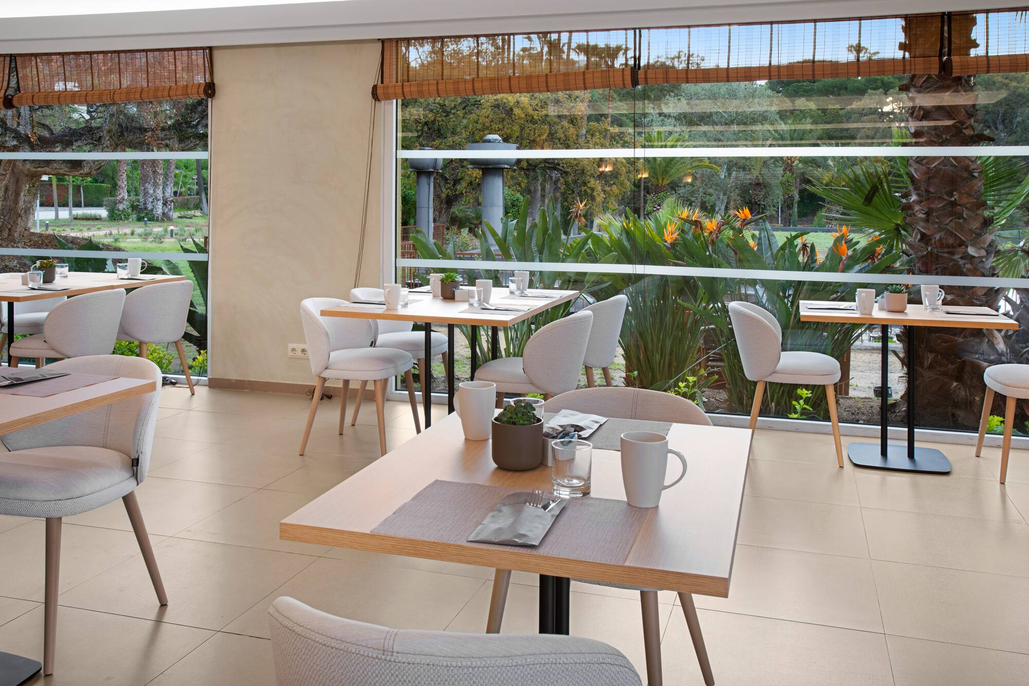 Modern restaurant with garden views, light wood tables, and grey chairs.