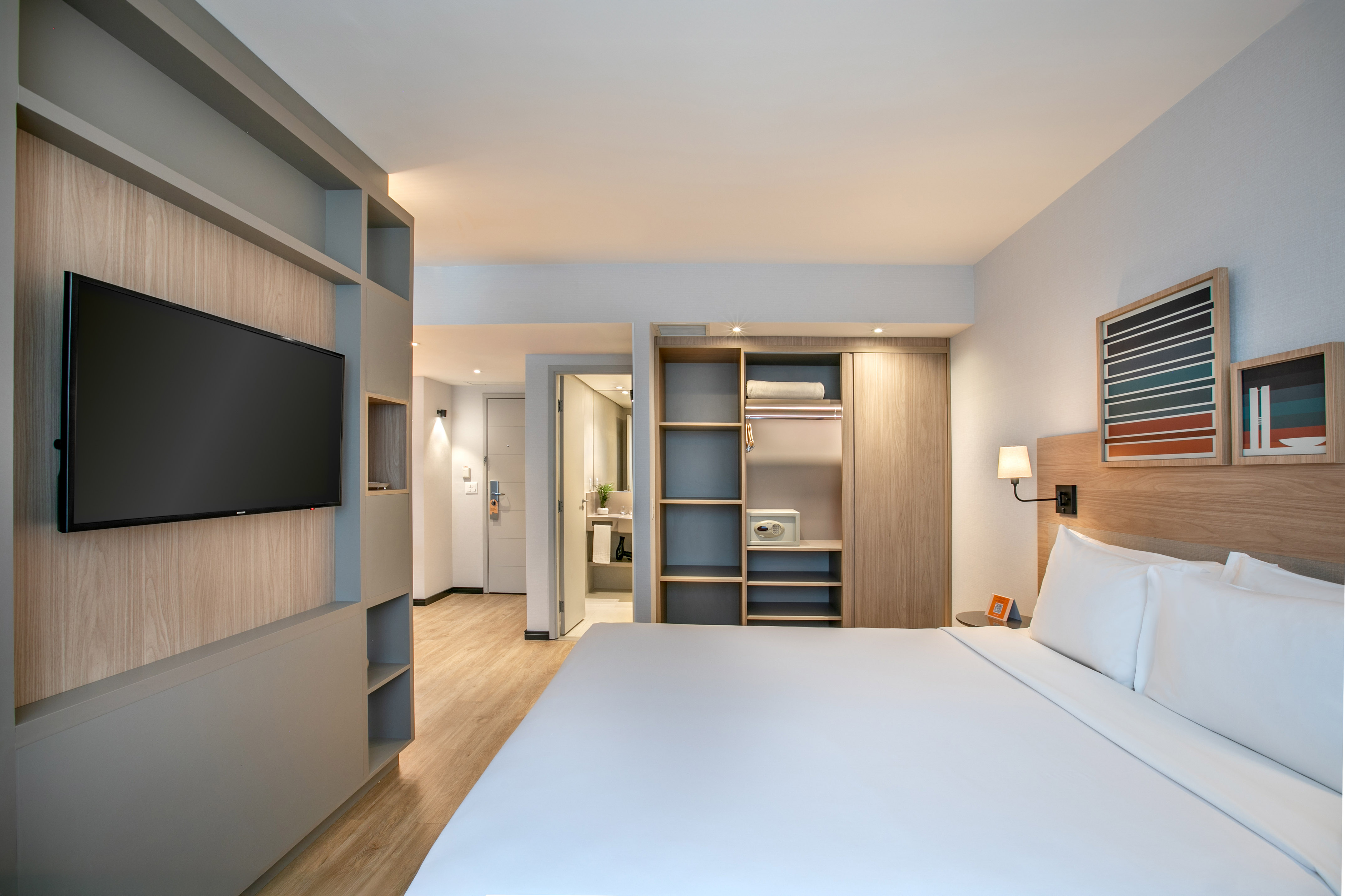 Modern hotel bedroom with white bed, wall-mounted TV, and integrated wood shelving.