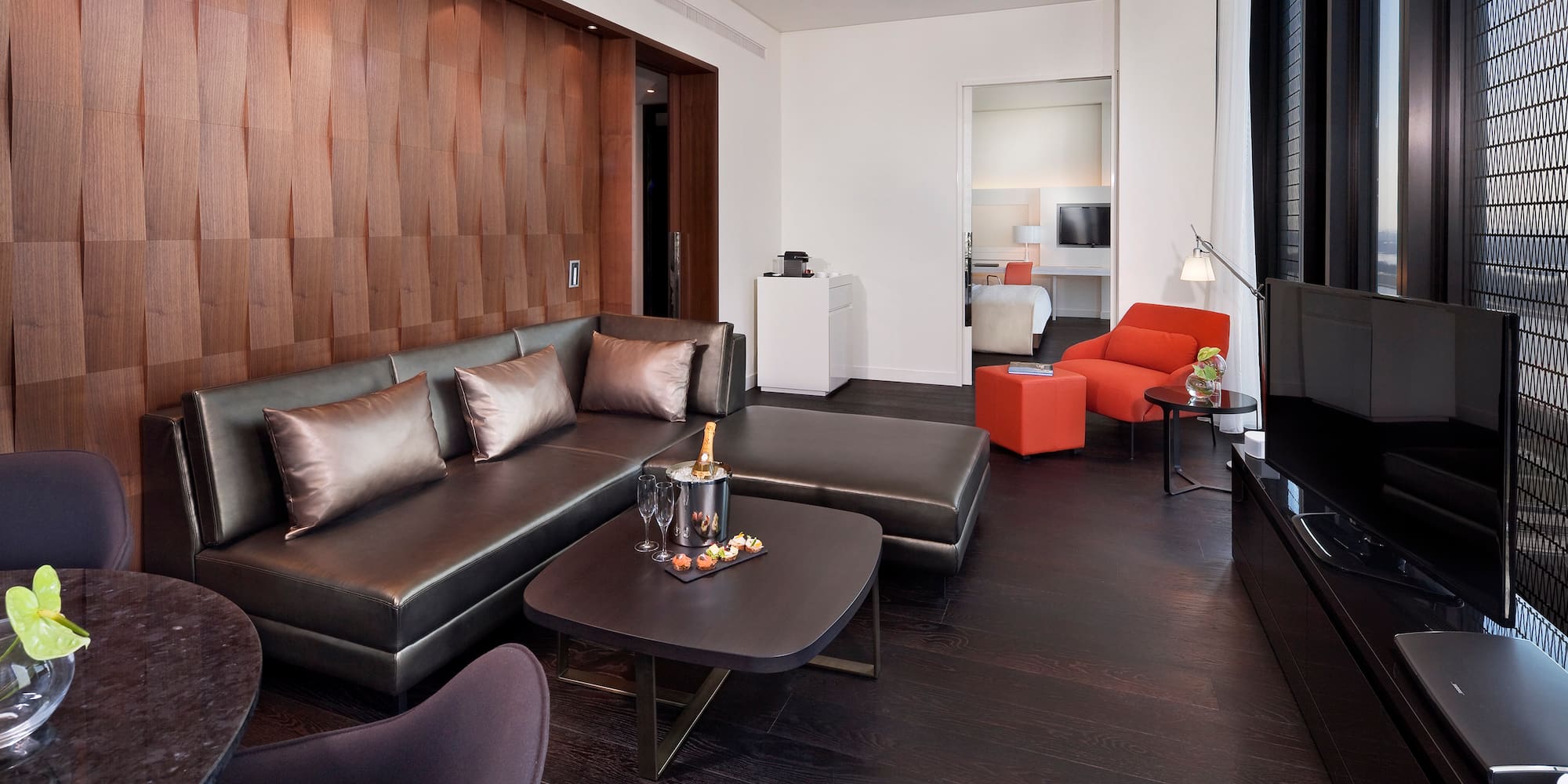Modern hotel suite with dark sofa, textured wood wall, orange chair, champagne.