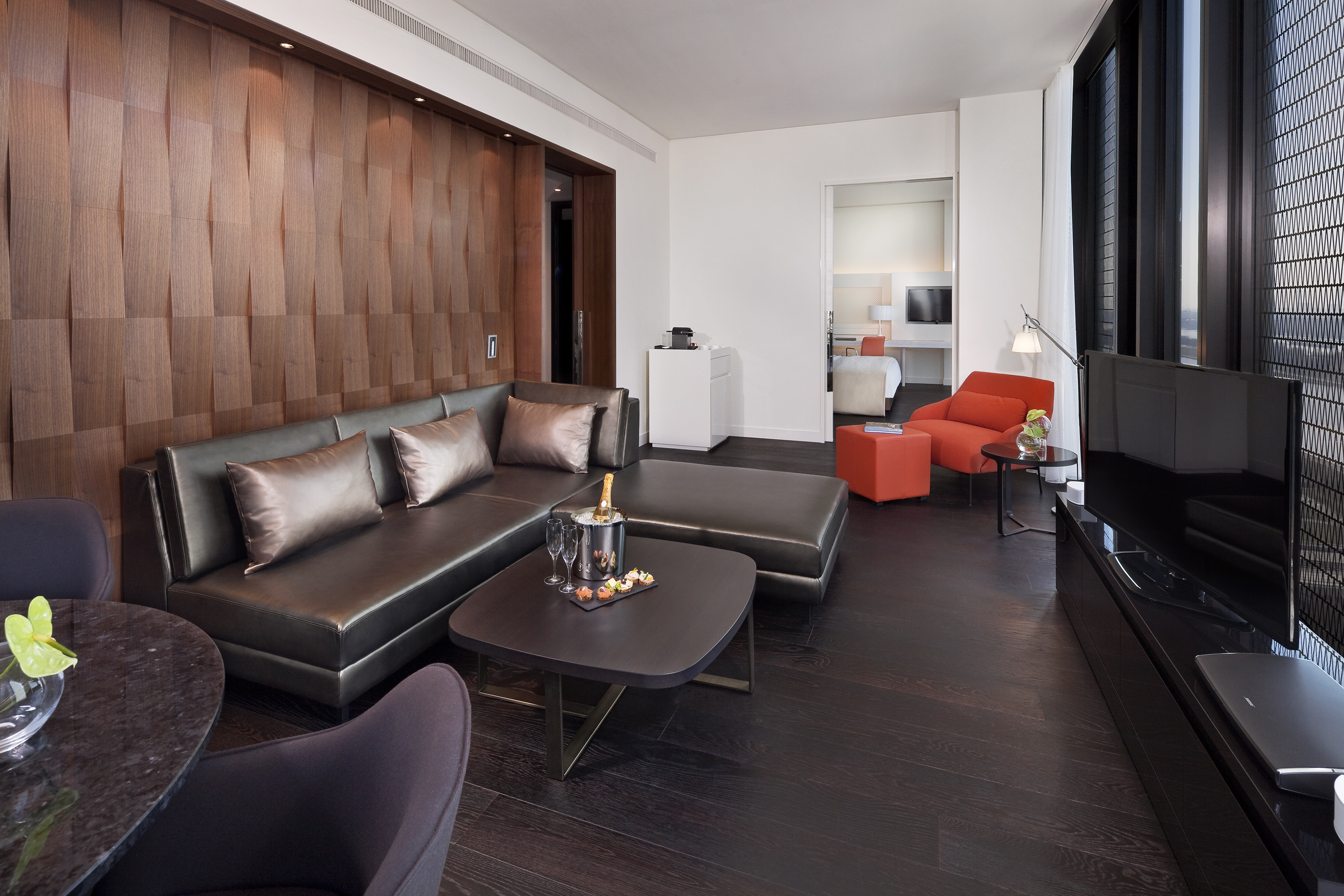 Modern hotel suite with dark sofa, textured wood wall, orange chair, champagne.
