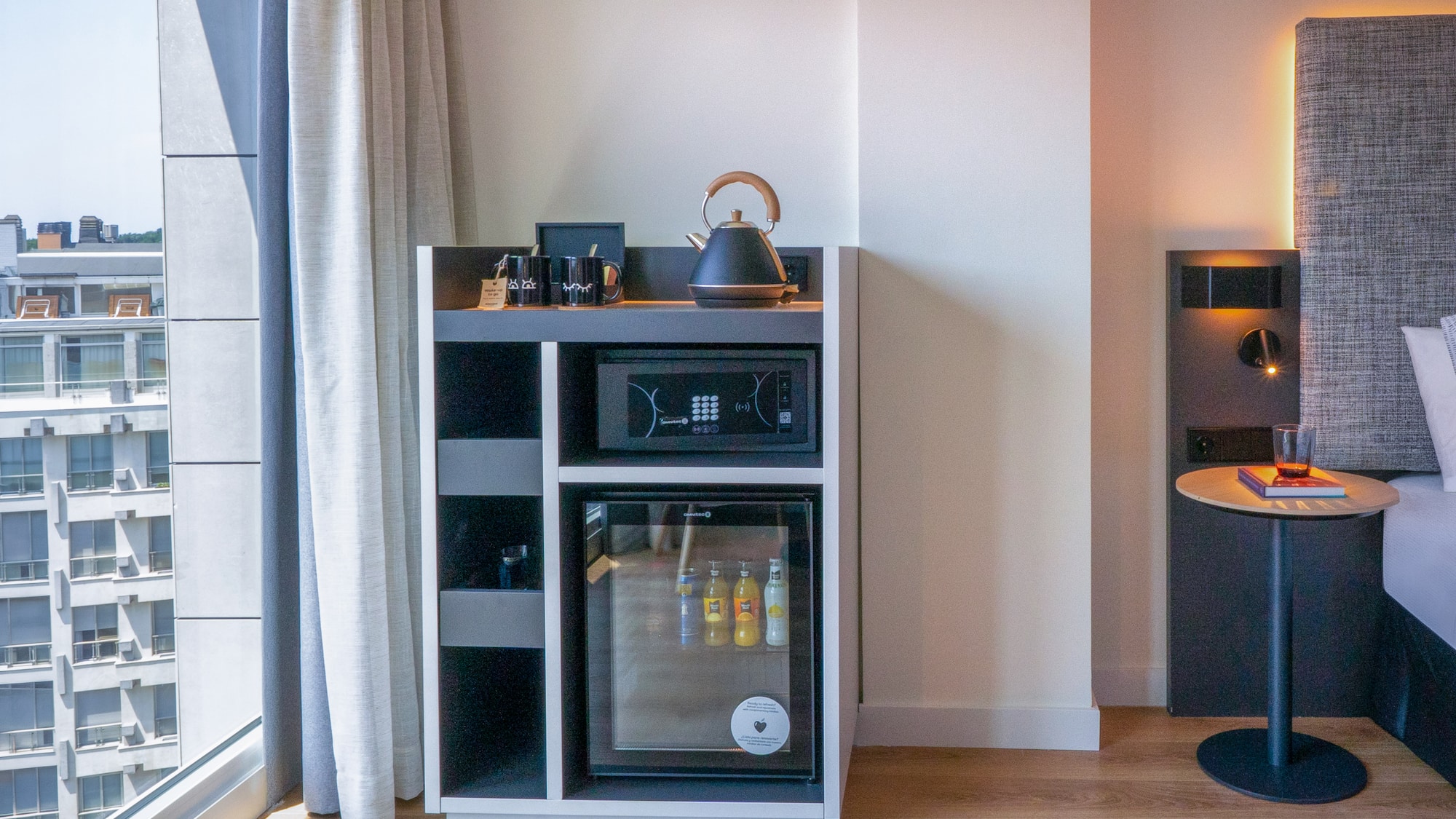 Habitación de hotel moderna con minibar, caja fuerte, hervidor y vista urbana.