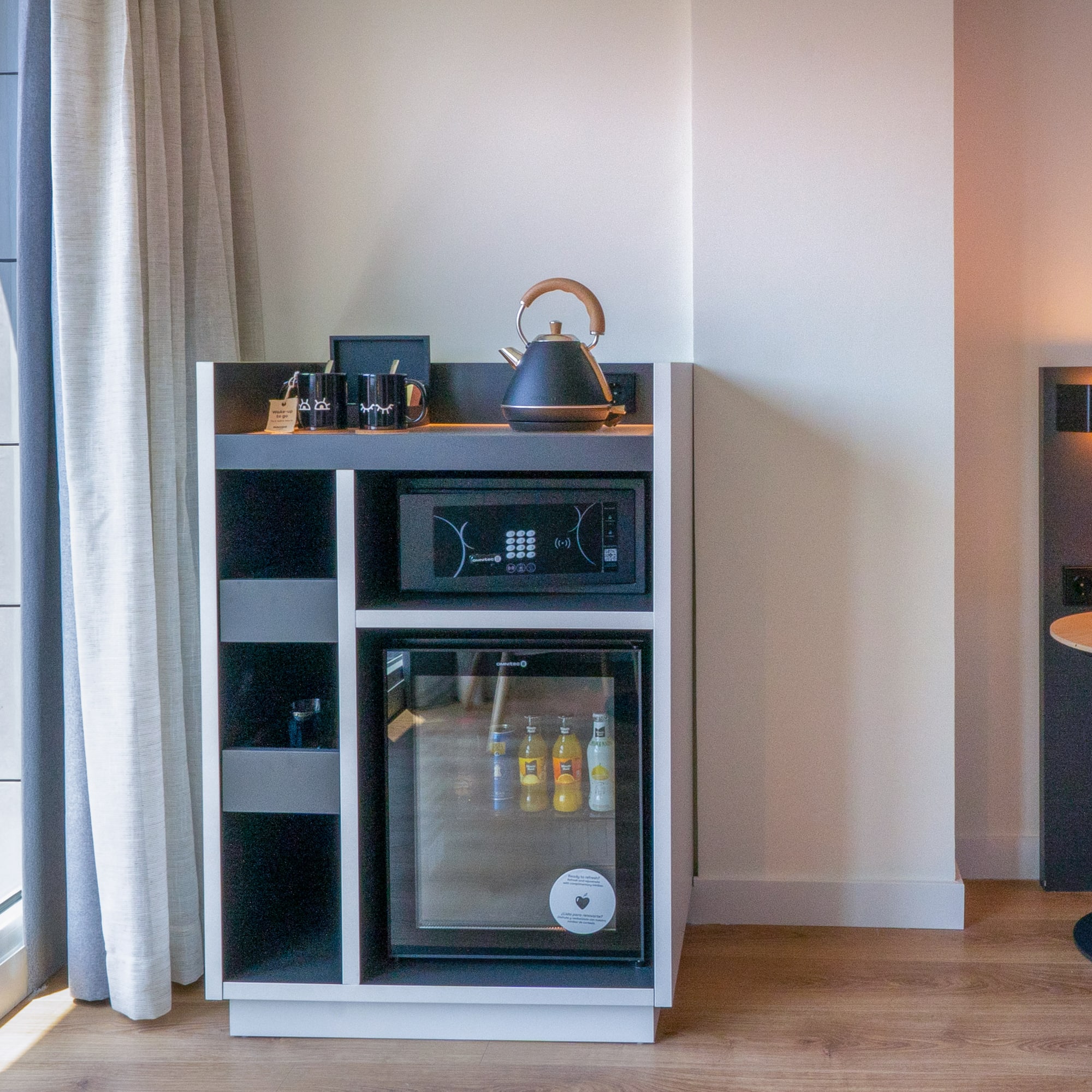Habitación de hotel moderna con minibar, caja fuerte, hervidor y vista urbana.