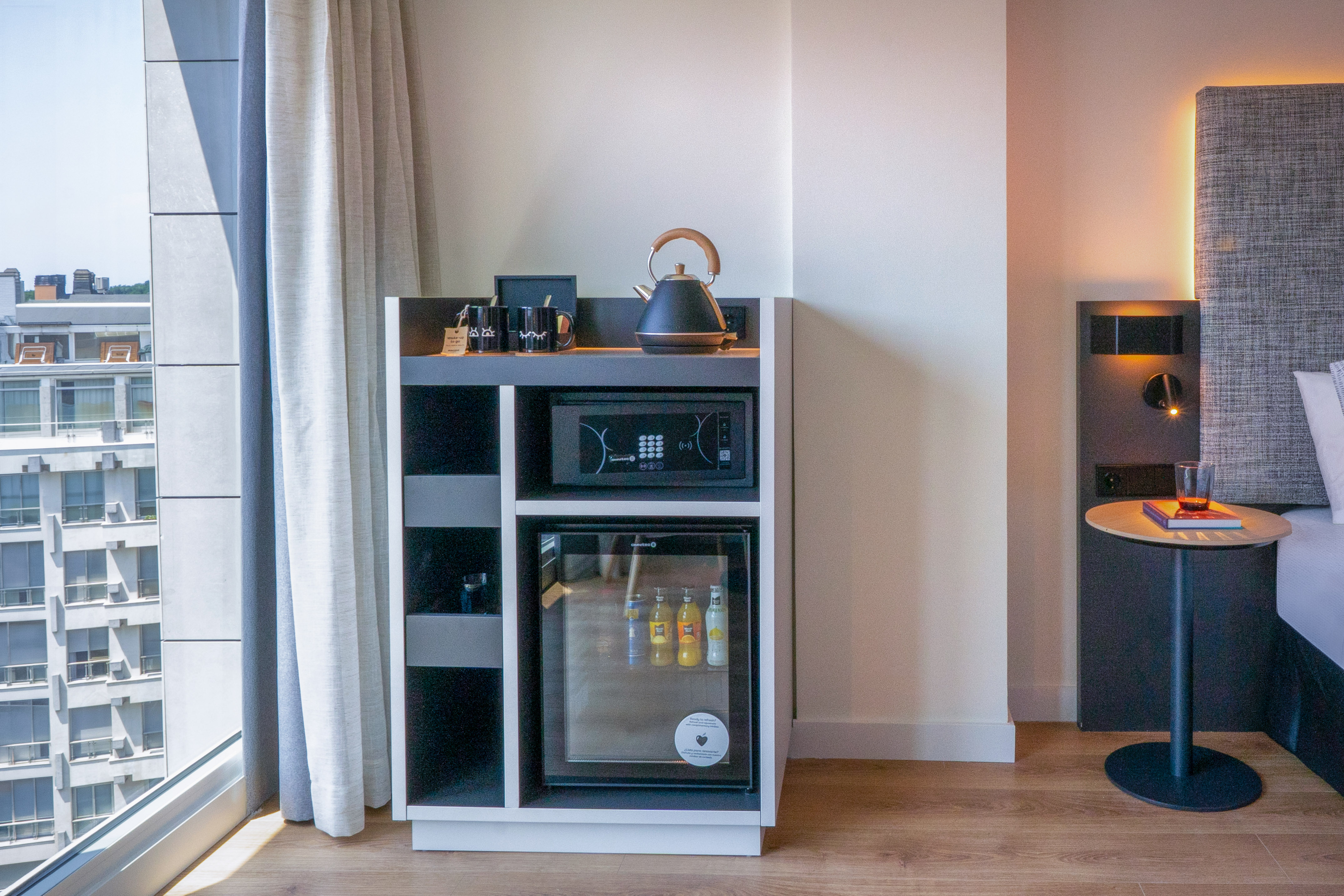 Habitación de hotel moderna con minibar, caja fuerte, hervidor y vista urbana.