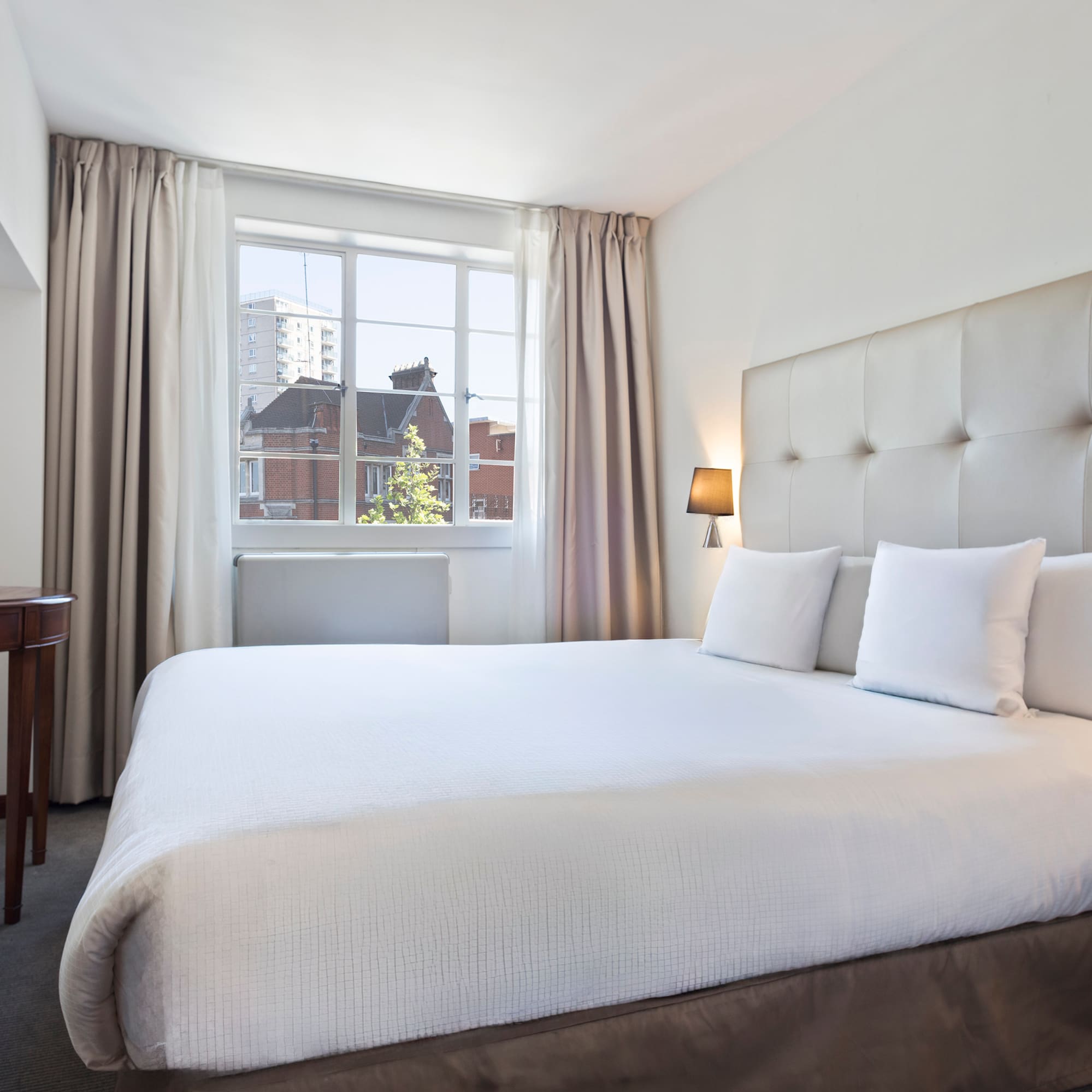 Bright hotel bedroom with large white bed, tufted headboard, and window view.