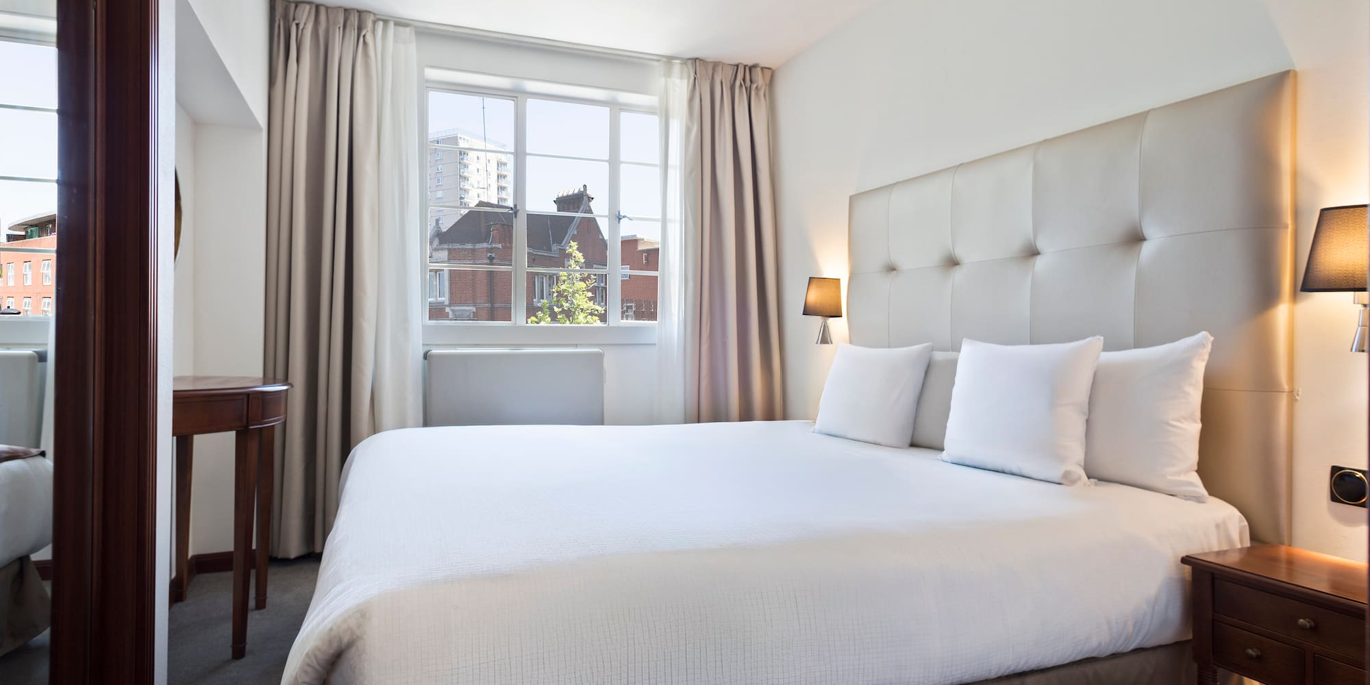 Bright hotel bedroom with white bed, upholstered headboard, and large window.