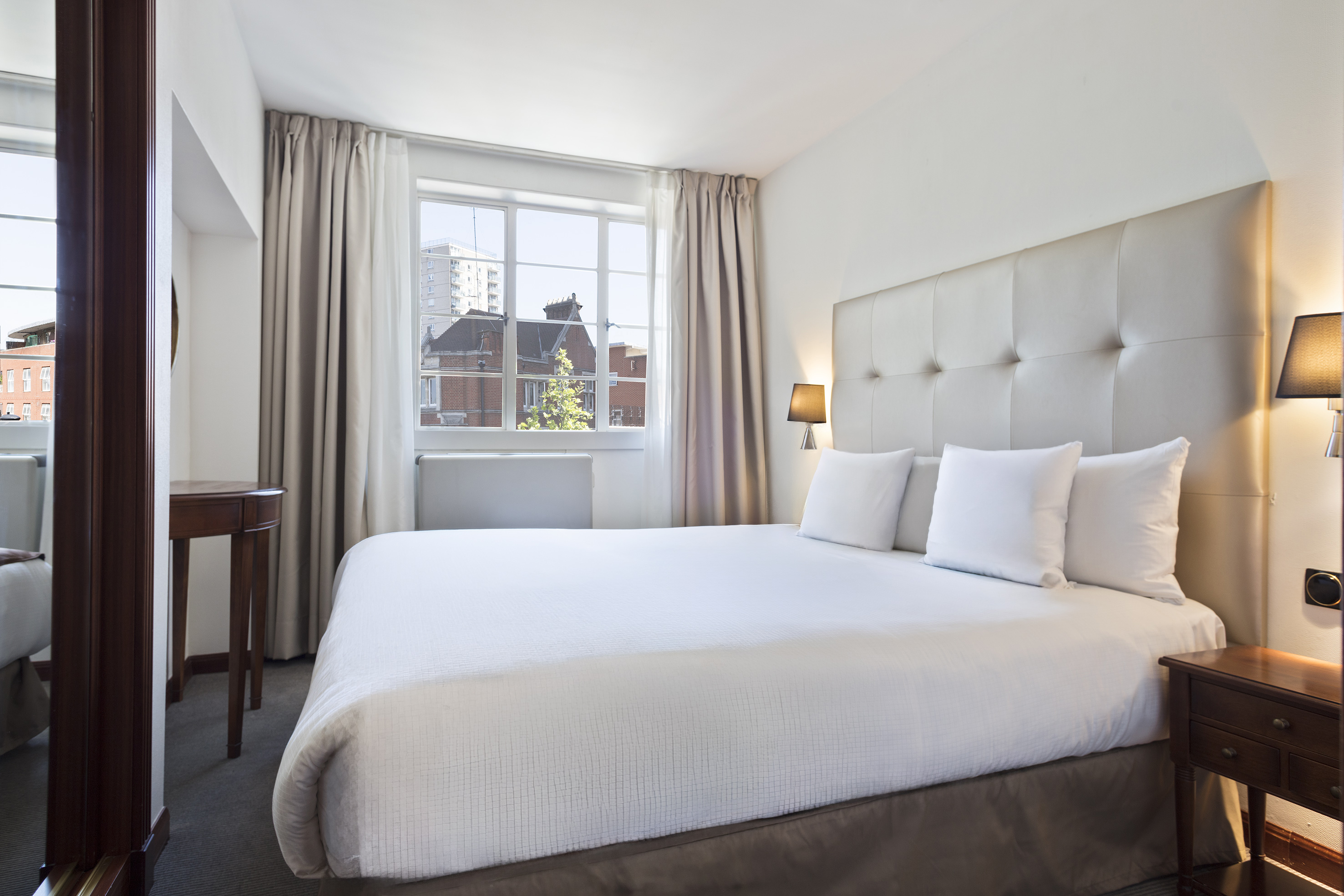 Modern hotel room with white bed, upholstered headboard, and sunlit window.