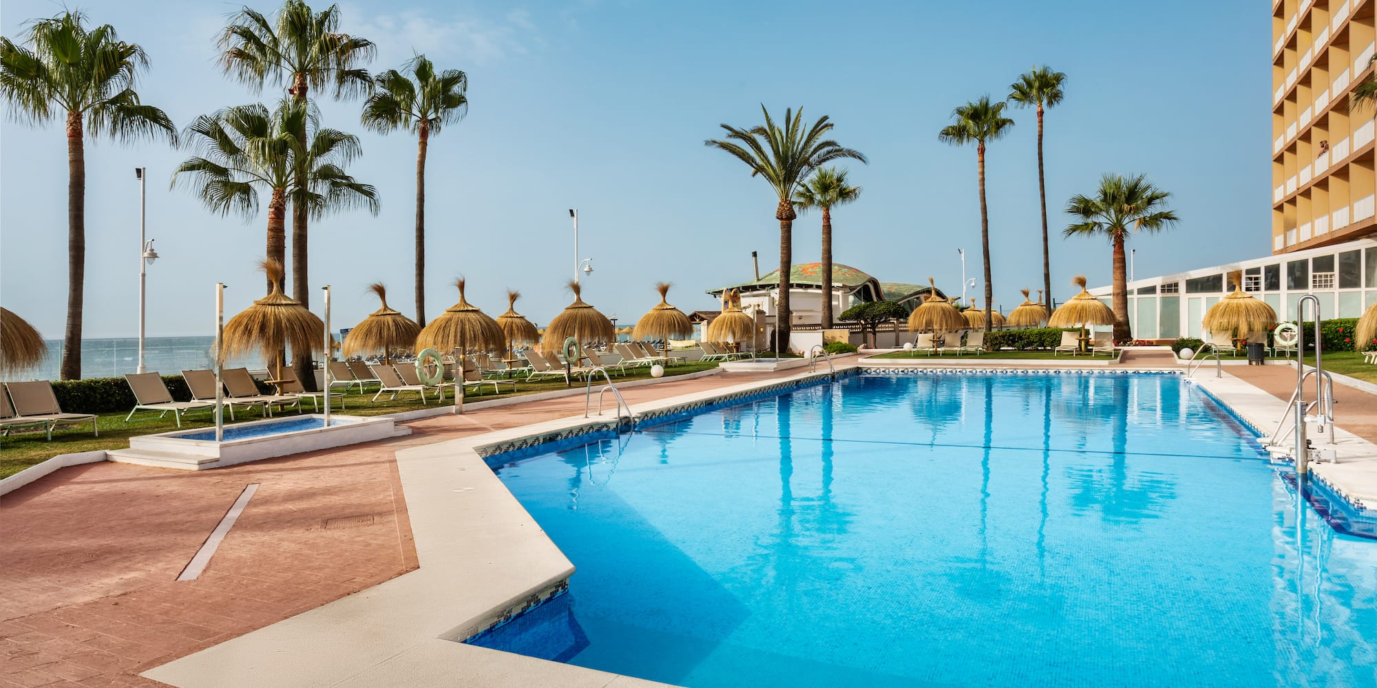 Sunny resort pool with palm trees, straw umbrellas, and hotel by the beach.