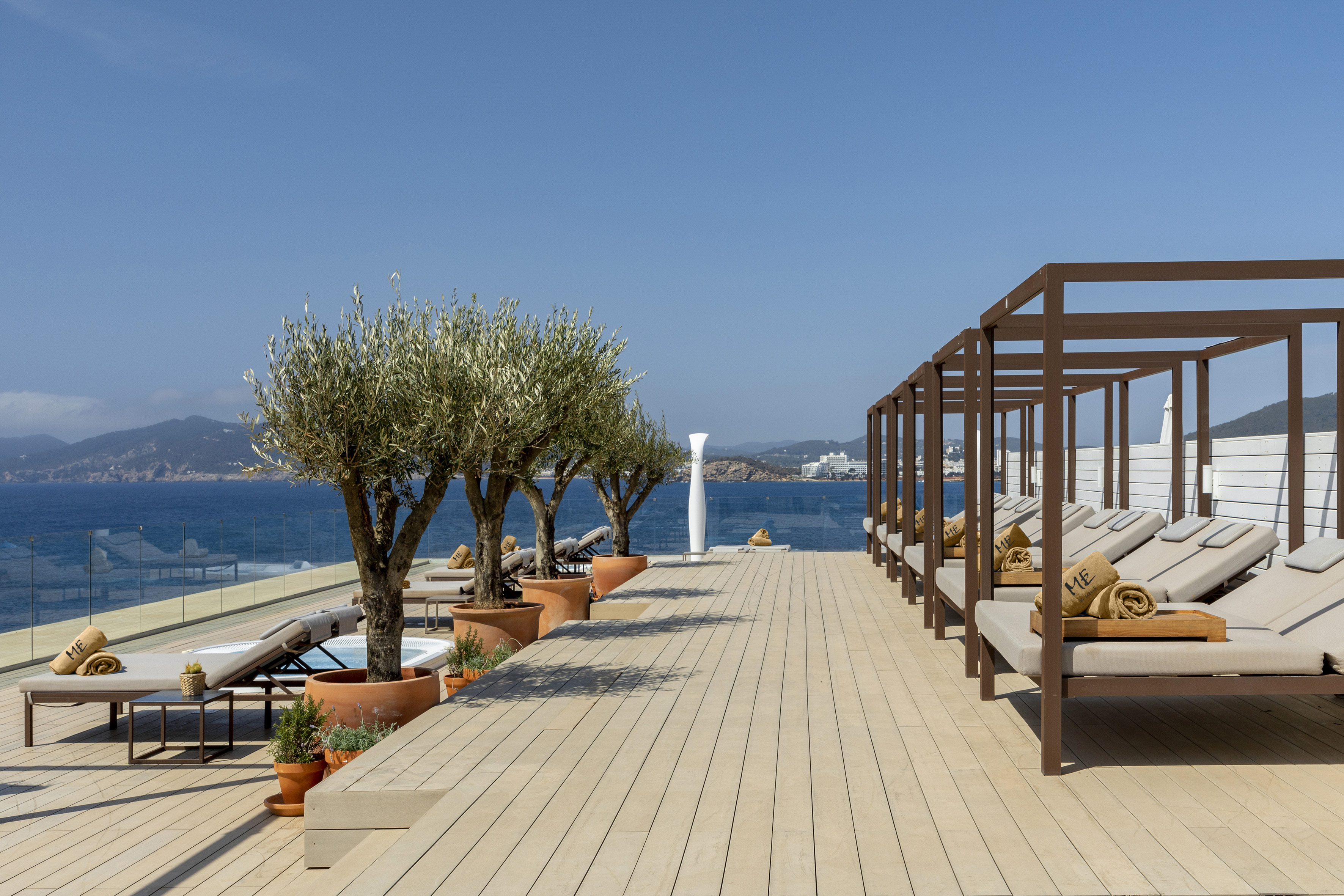Mediterranean resort deck with ocean view, stylish daybeds, and olive trees.