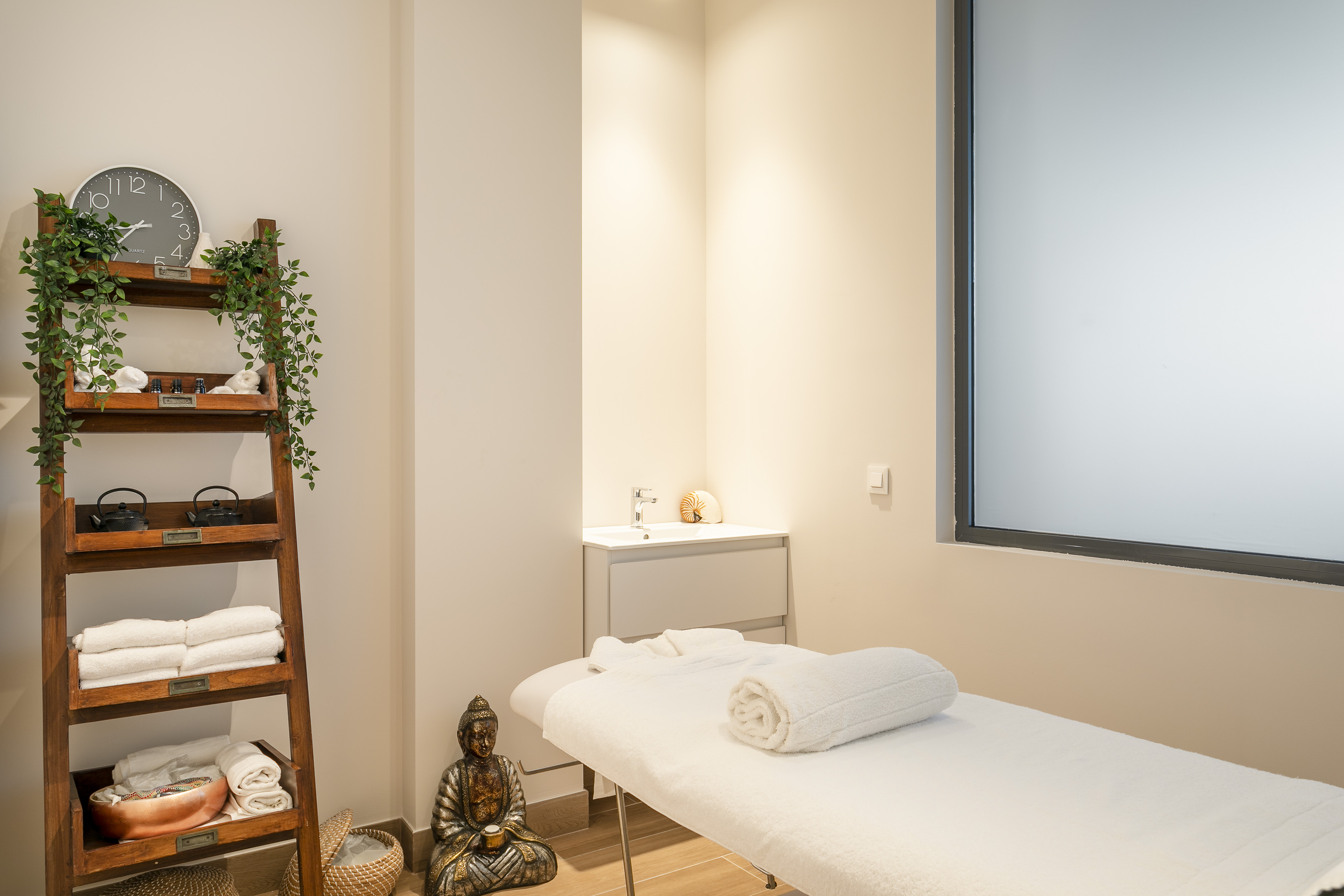 Modern spa therapy room with massage bed, Buddha, and wooden shelves.
